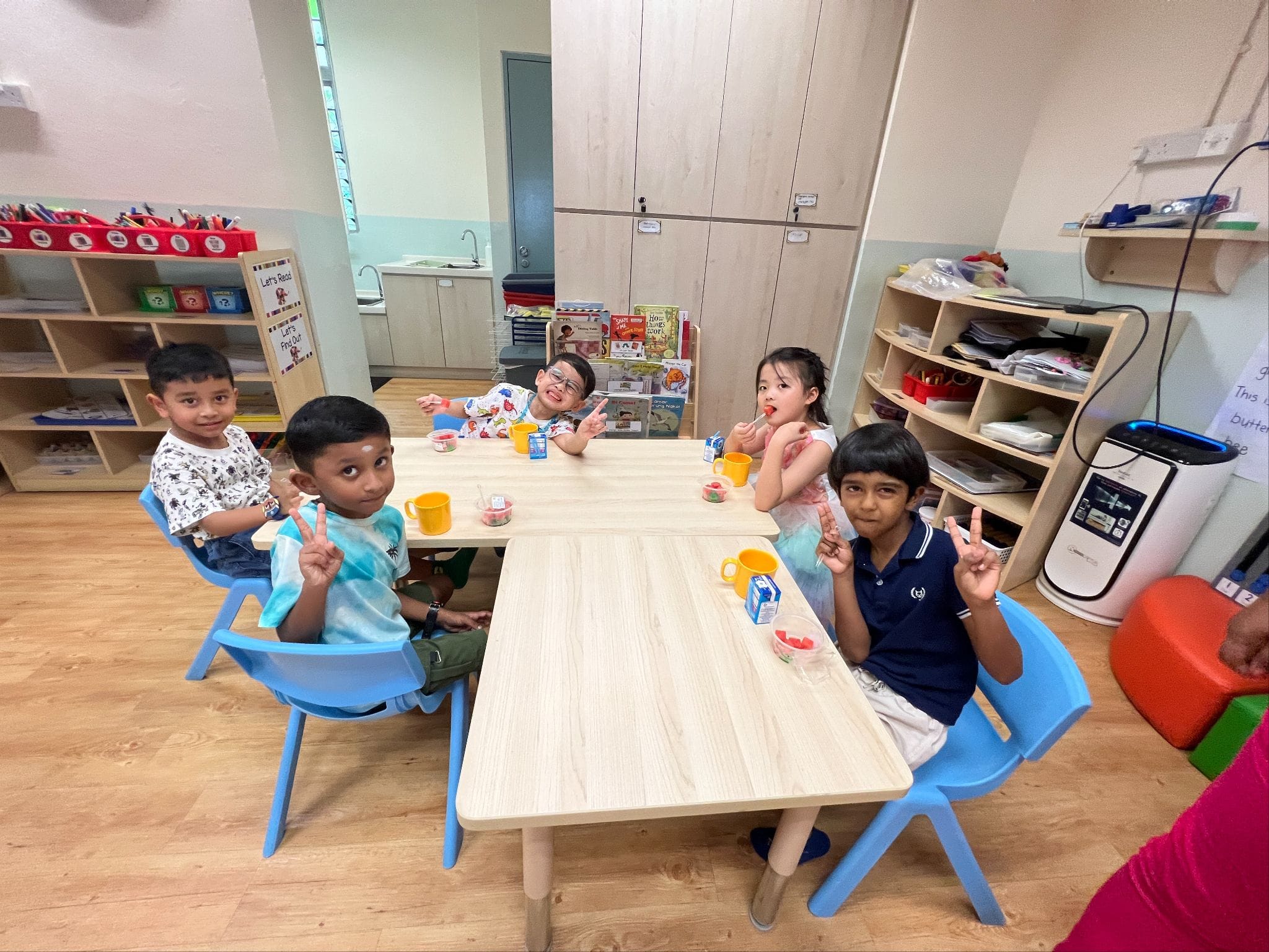 Five young children sit at tables in a classroom, smiling and making peace signs with snacks and juice boxes.