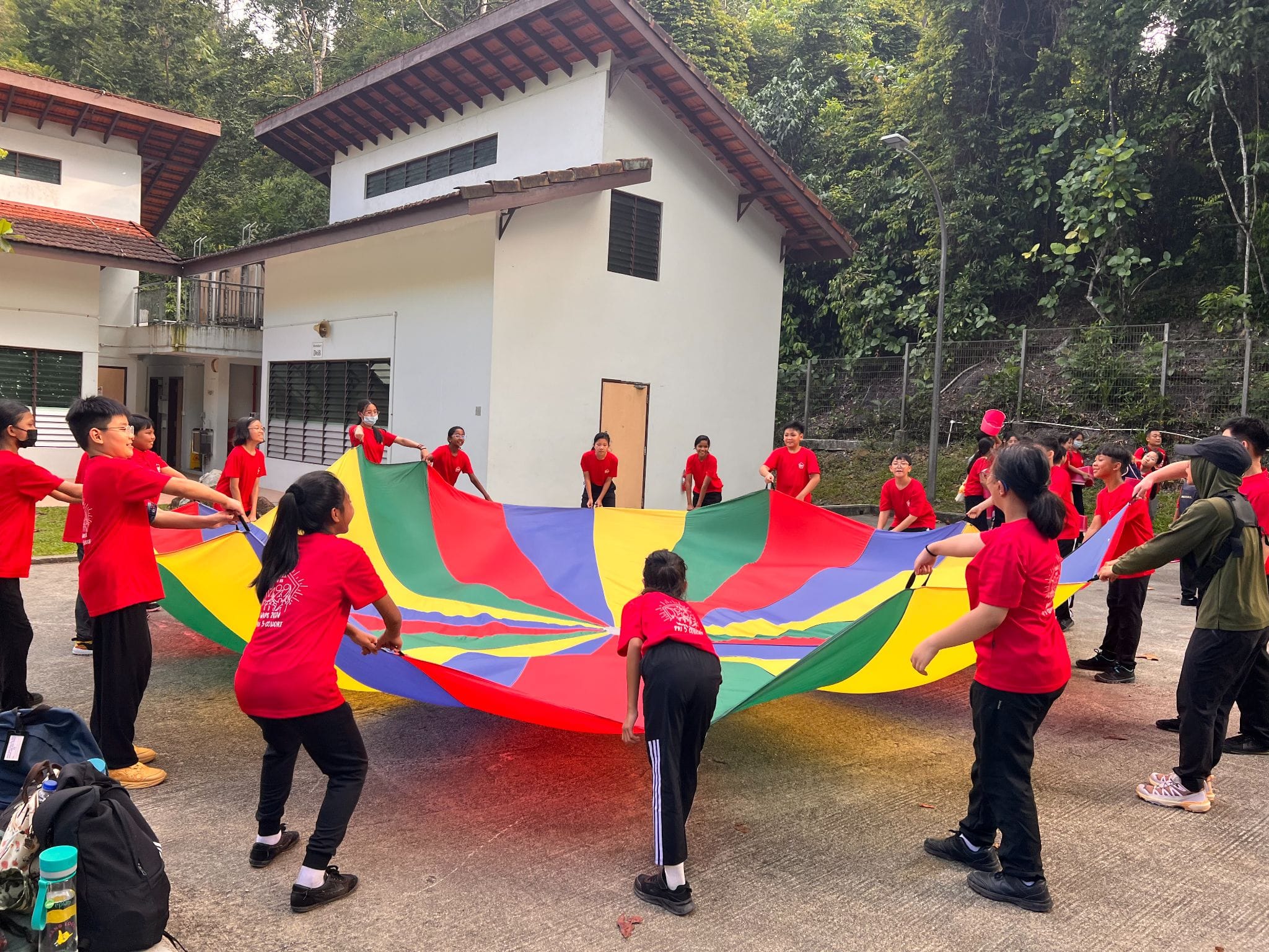 Children in red shirts stand in a circle outdoors, holding the edges of a large colorful parachute.