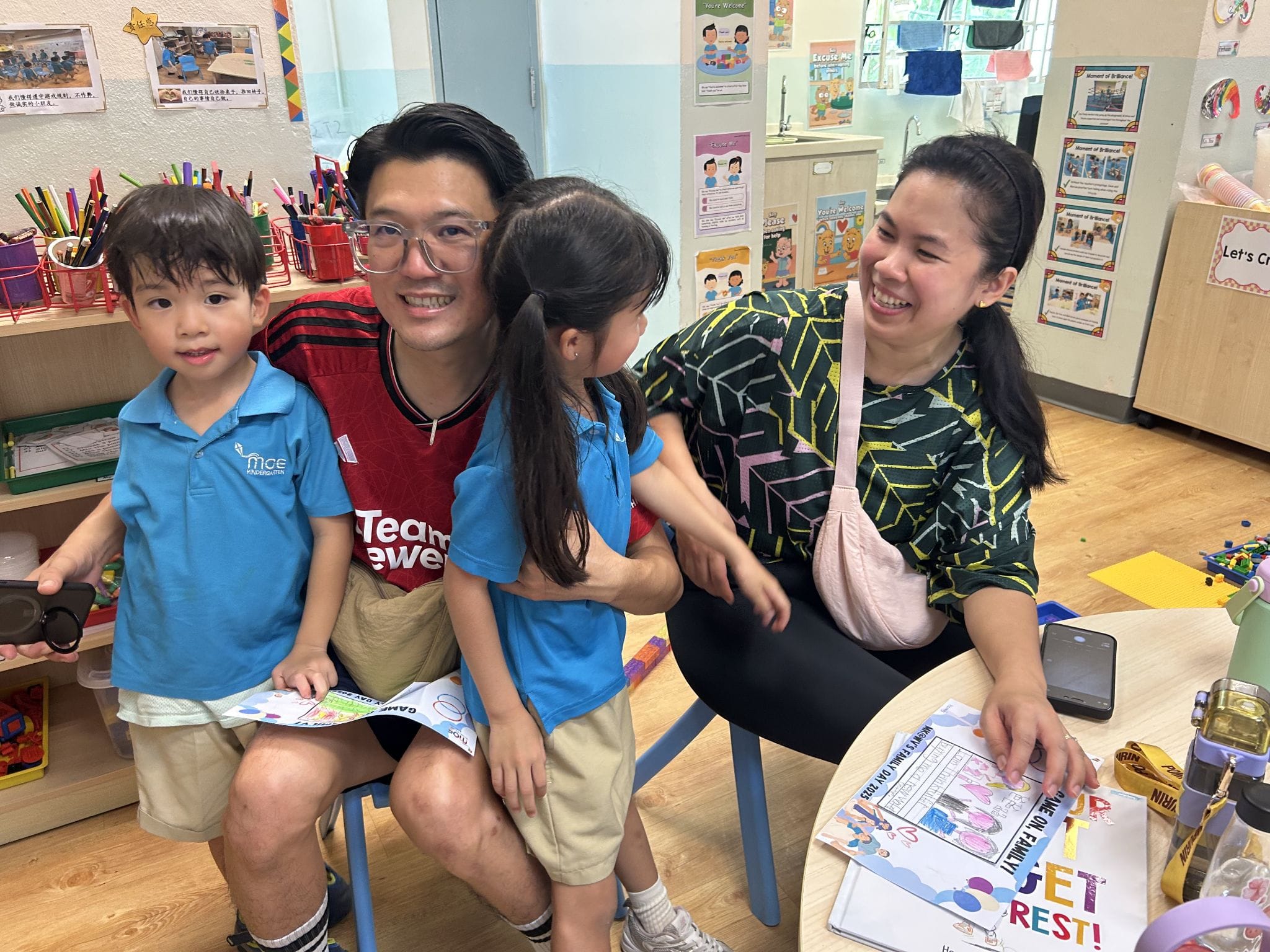 A smiling family of four, two adults and two young children in blue uniforms, sitting together in a colorful classroom.
