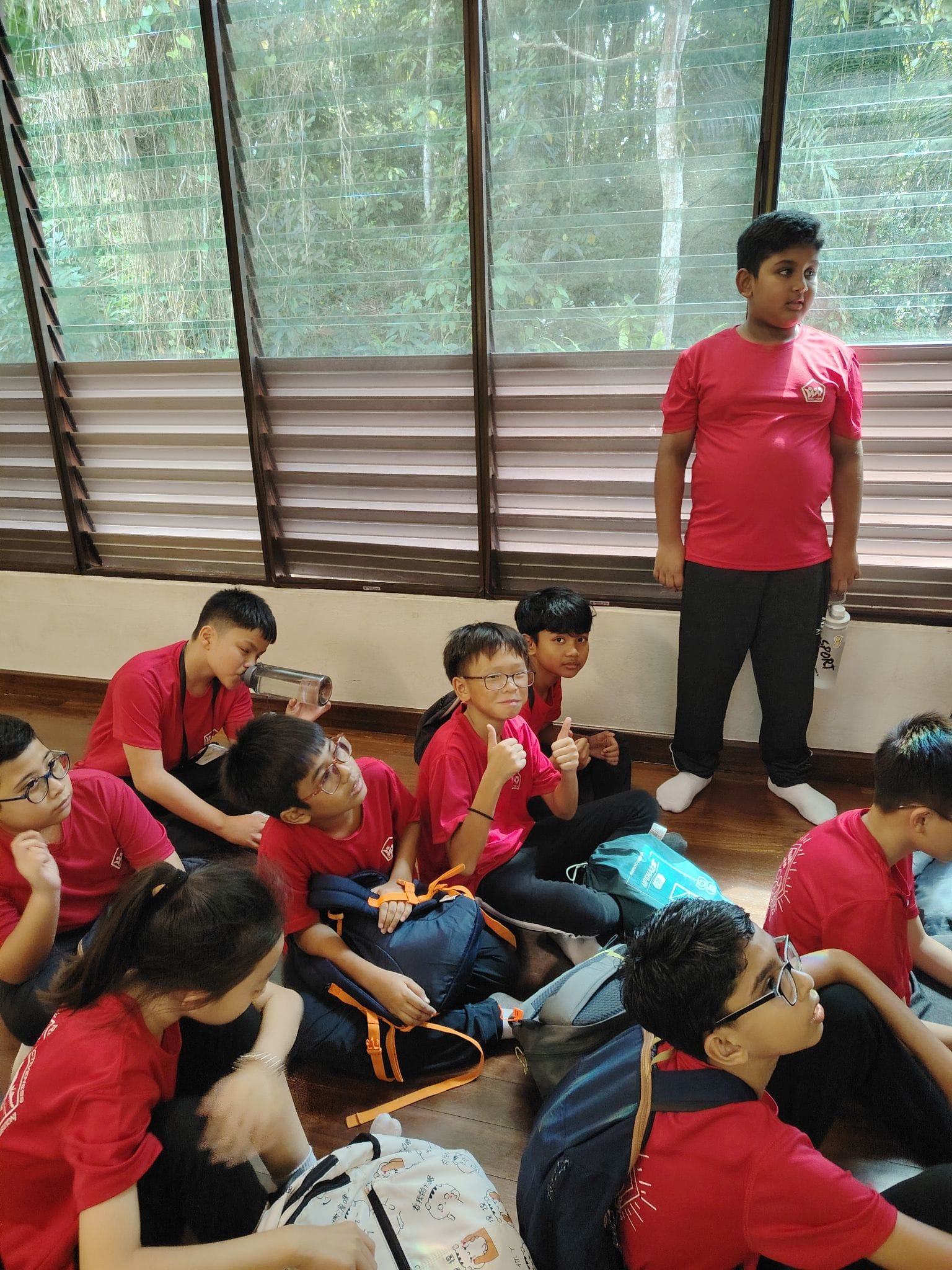 A group of children in red shirts sit on a floor with backpacks while one boy stands near a window giving thumbs up.