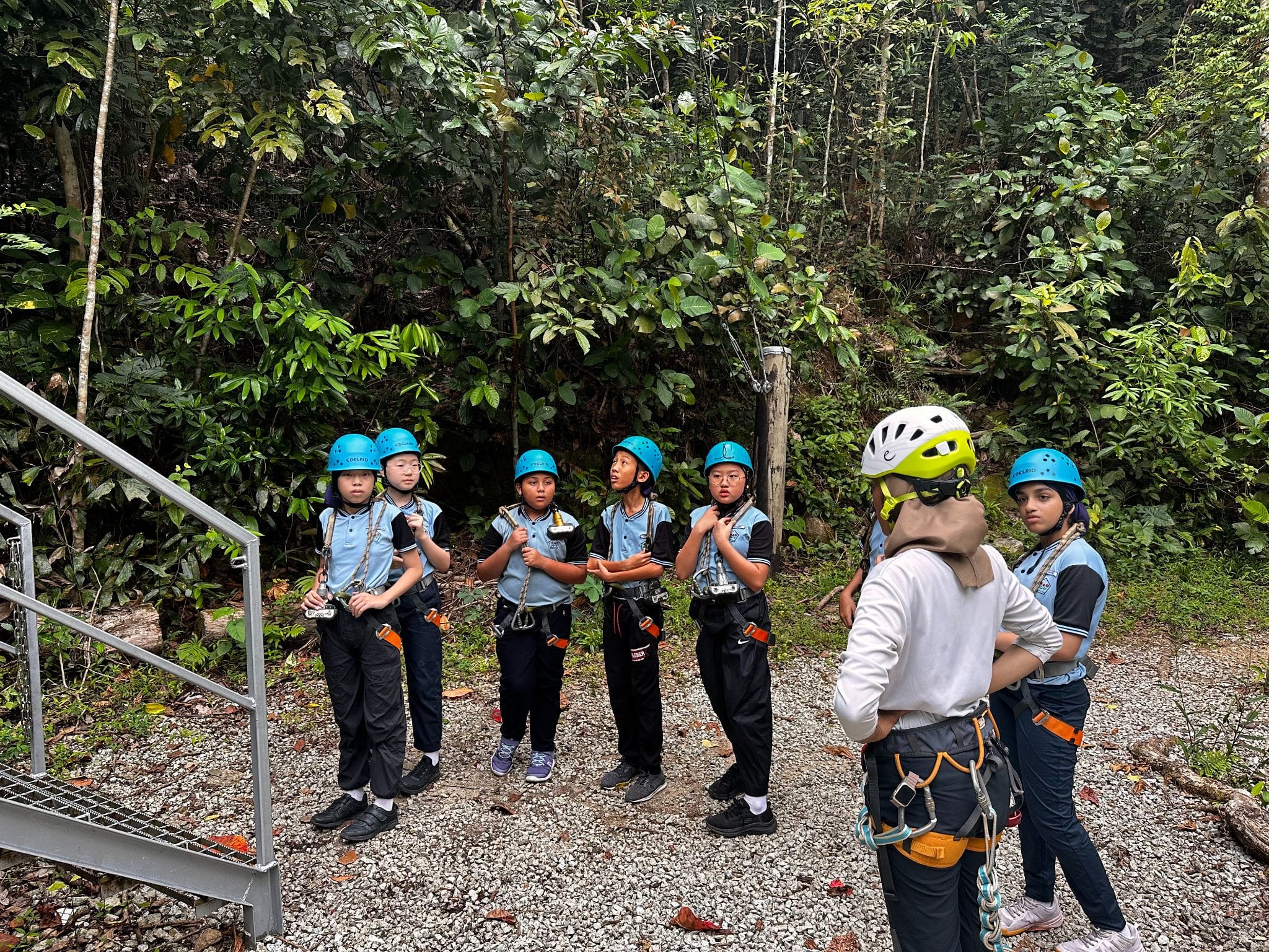 A group of children in blue helmets and harnesses listen to an instructor in a forest setting.