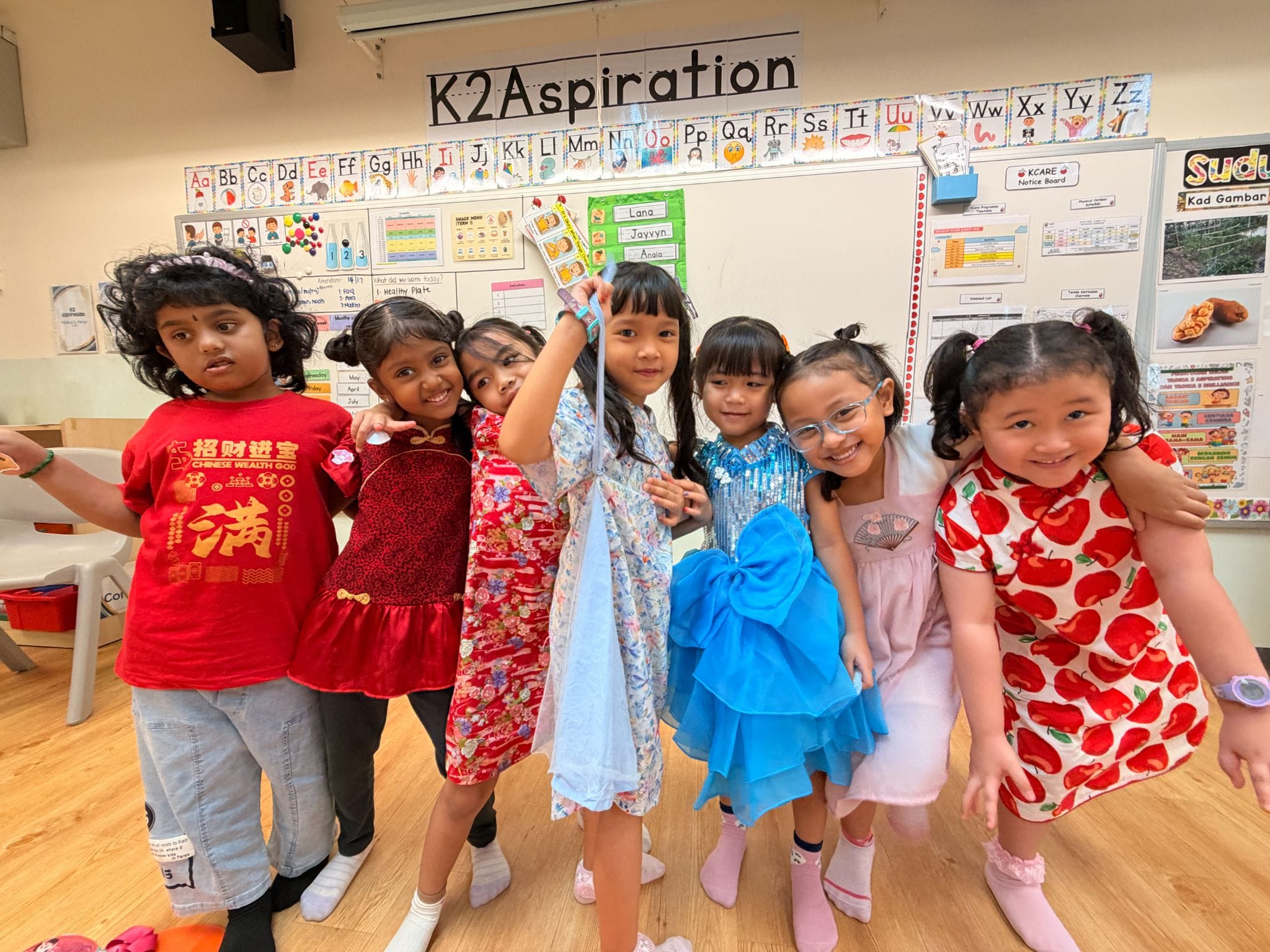 Seven young girls in colorful outfits pose together smiling in a classroom labeled K2 Aspiration.