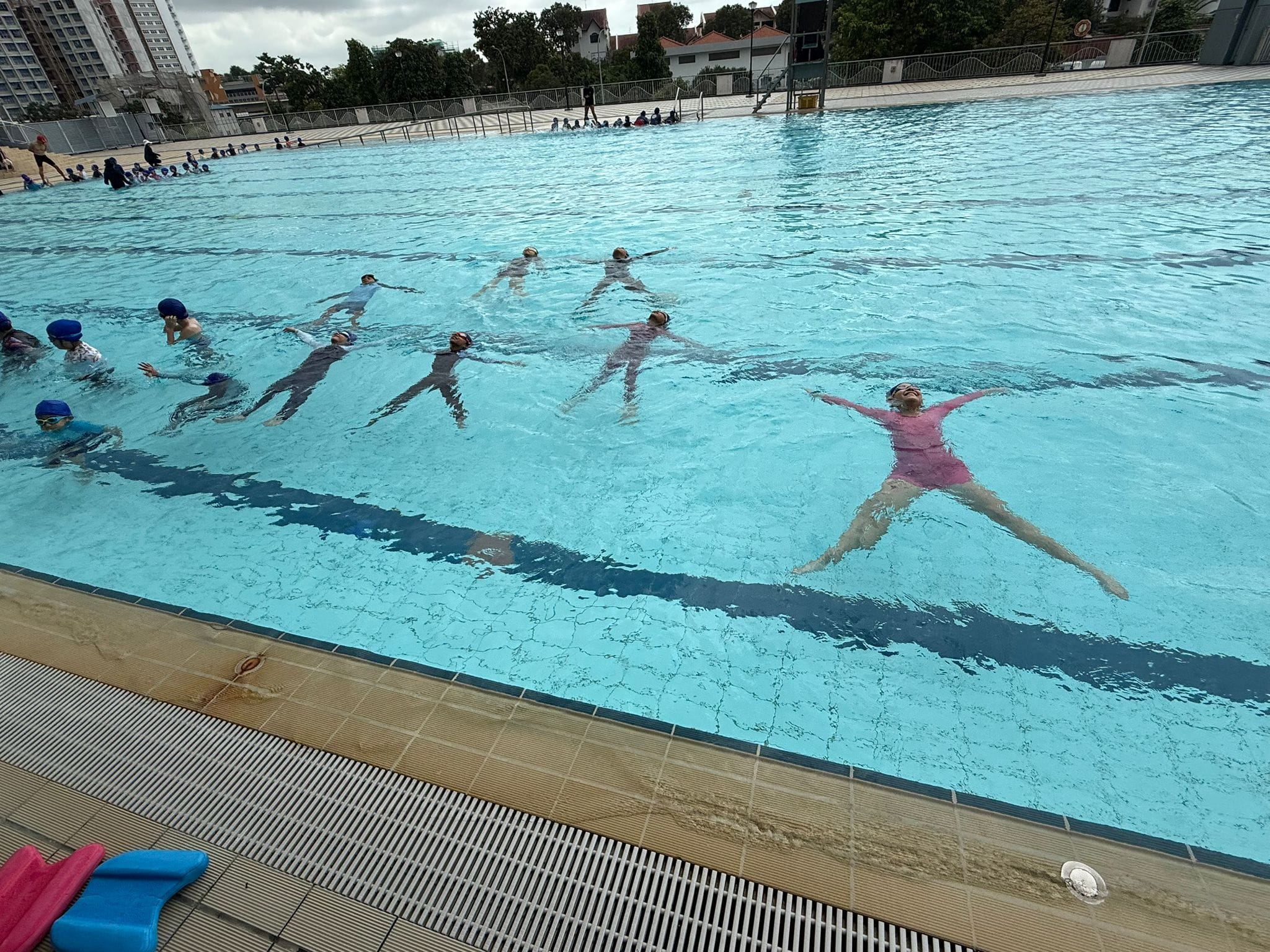 Several children float on their backs in an outdoor pool during a swimming lesson, with more students waiting at the edge.