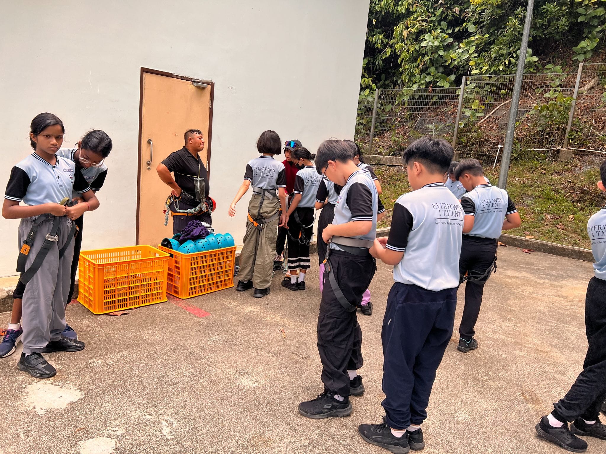 Students in light blue school uniforms put on climbing harnesses outdoors, guided by an instructor near orange crates with helmets.