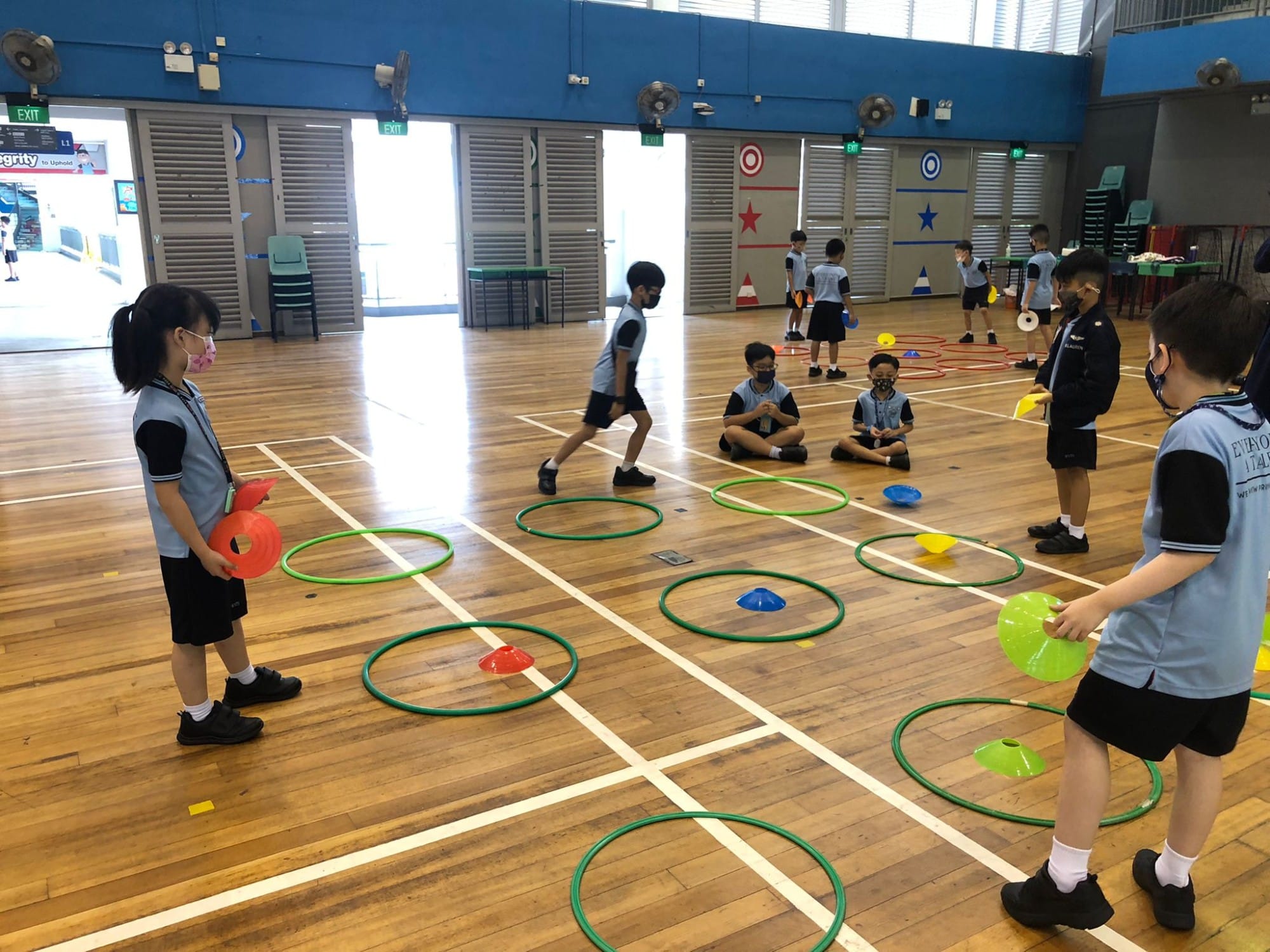 Children in school uniforms and masks do a PE activity with green hoops and colorful cones on a wooden gym floor.
