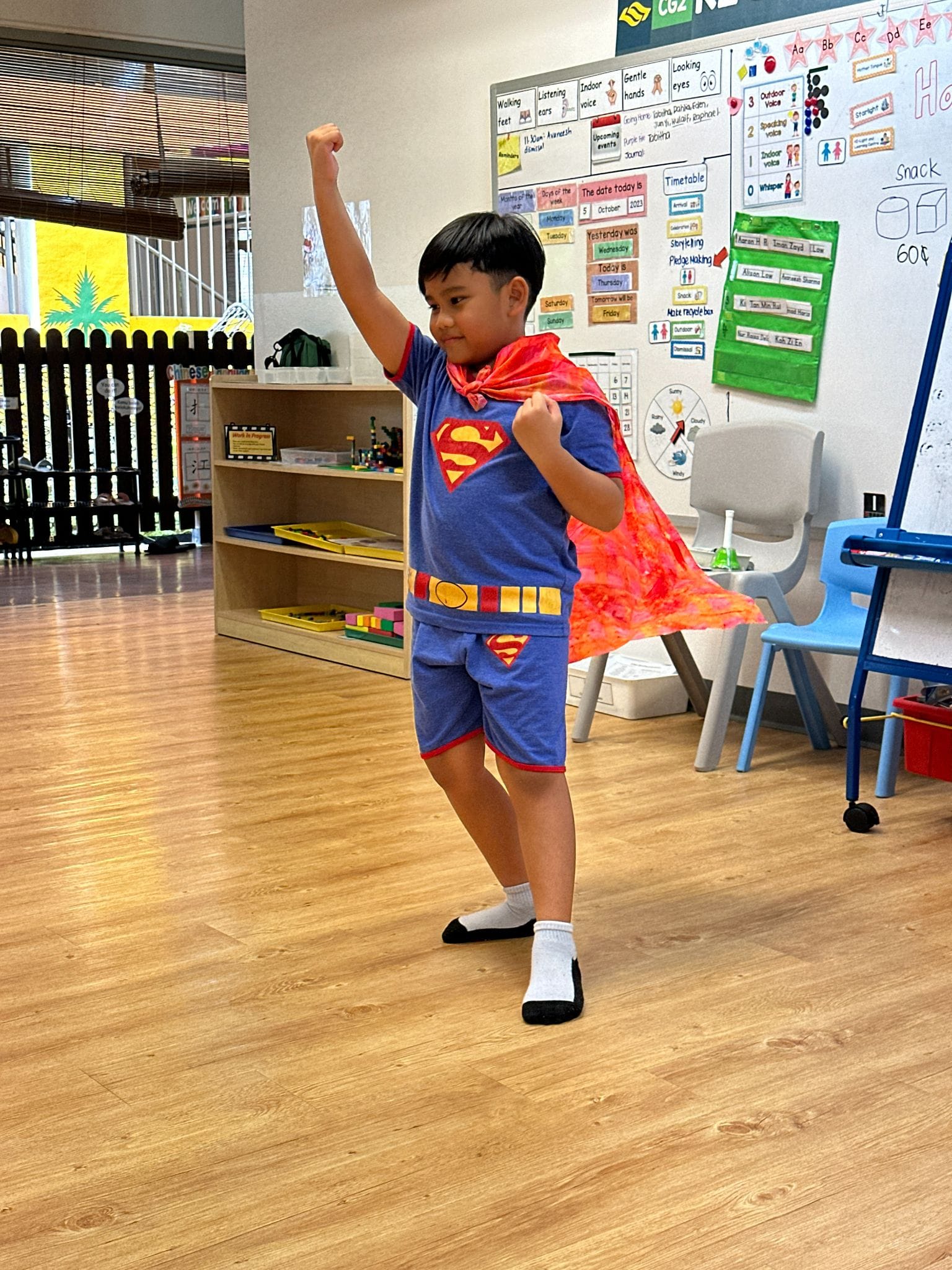 Young boy in a Superman costume with a red cape poses with fist raised in a classroom.