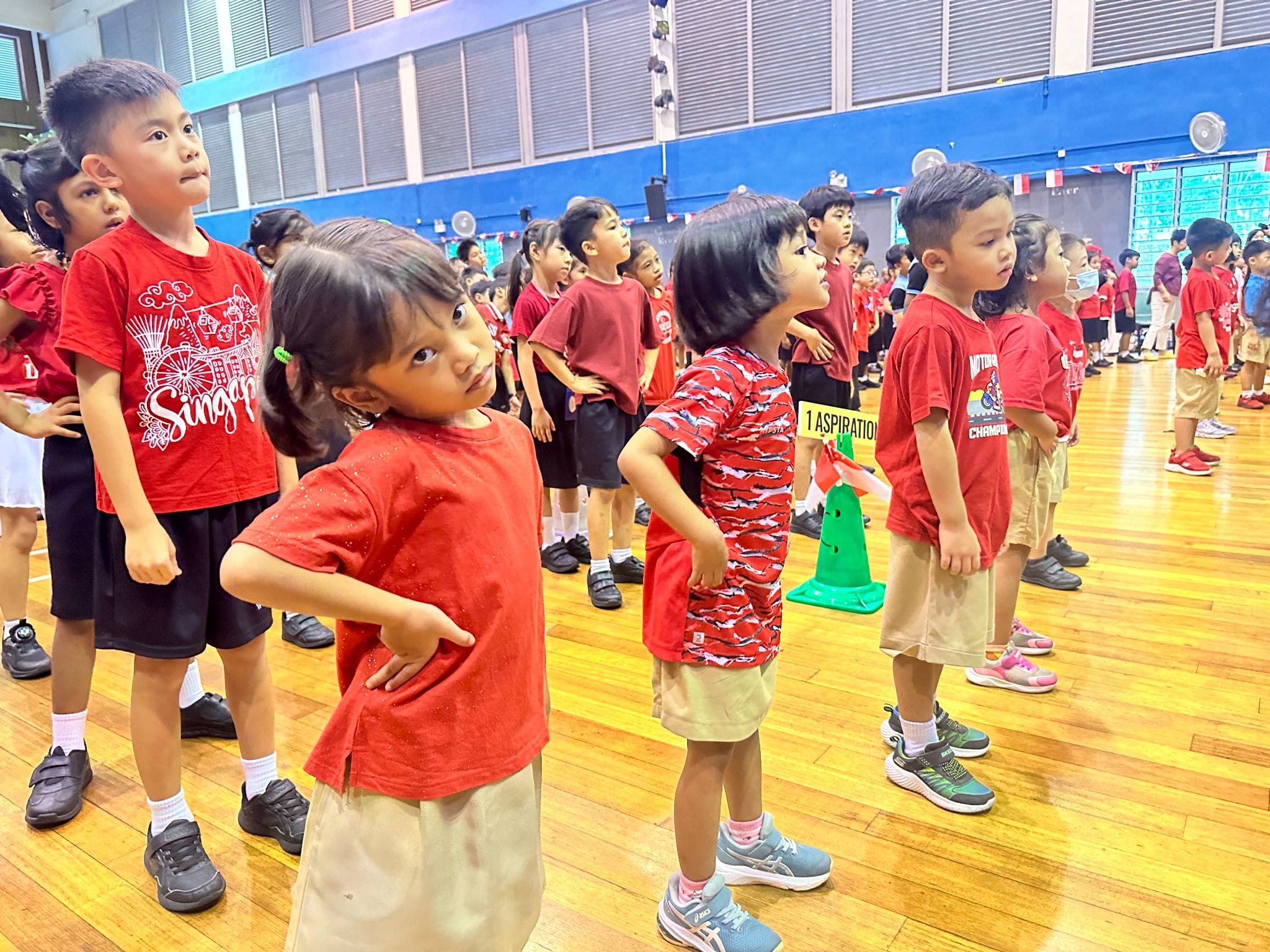 Young children in red shirts stand in rows on a wooden gym floor during a school assembly event.
