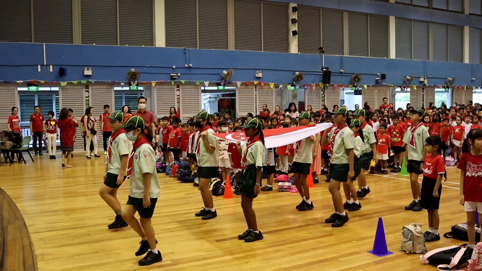 Children in scout uniforms carry the Singapore flag during a school ceremony in an indoor hall.
