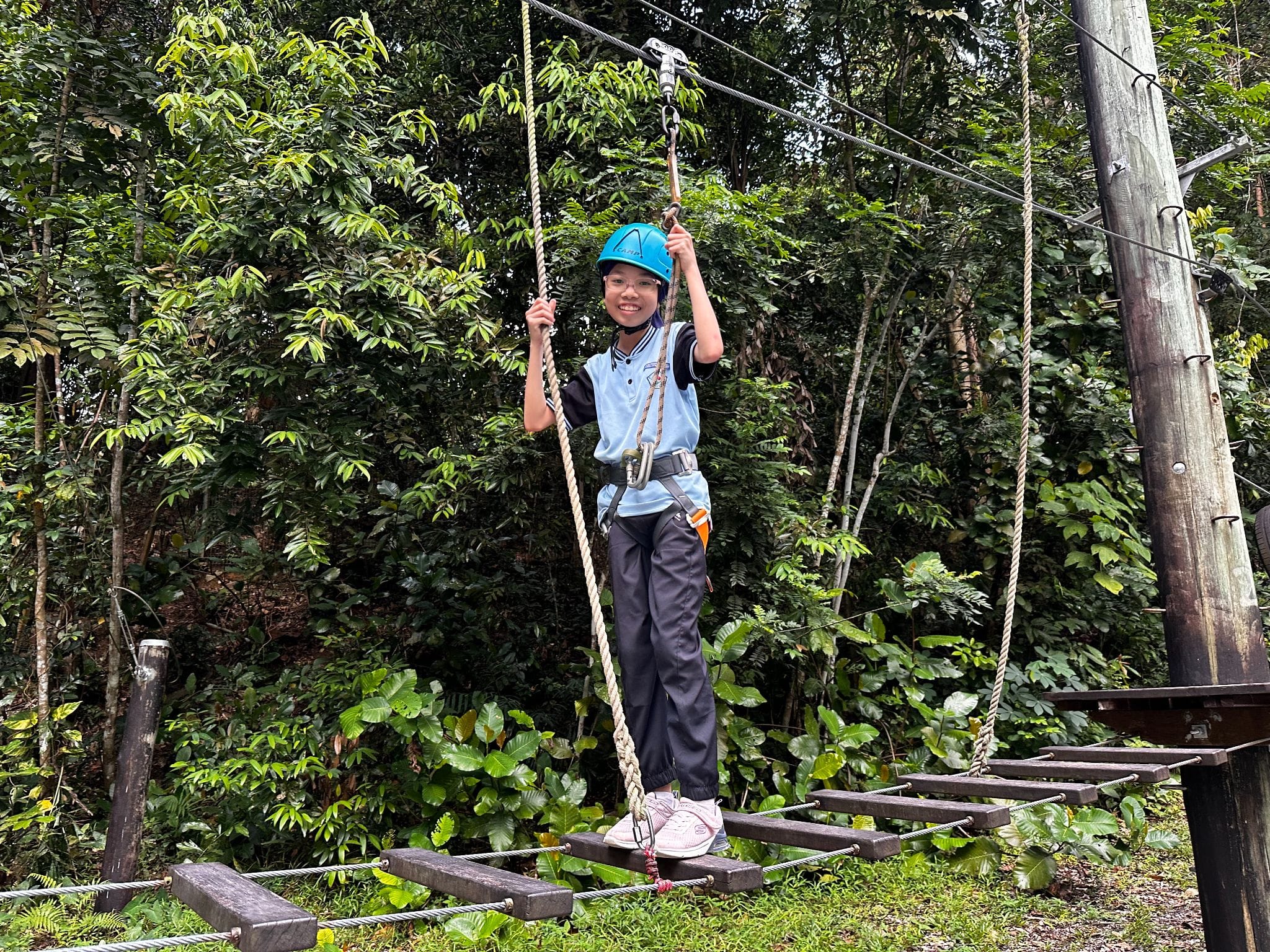 A smiling child in a blue helmet and harness stands on a rope bridge at an outdoor adventure course surrounded by trees.