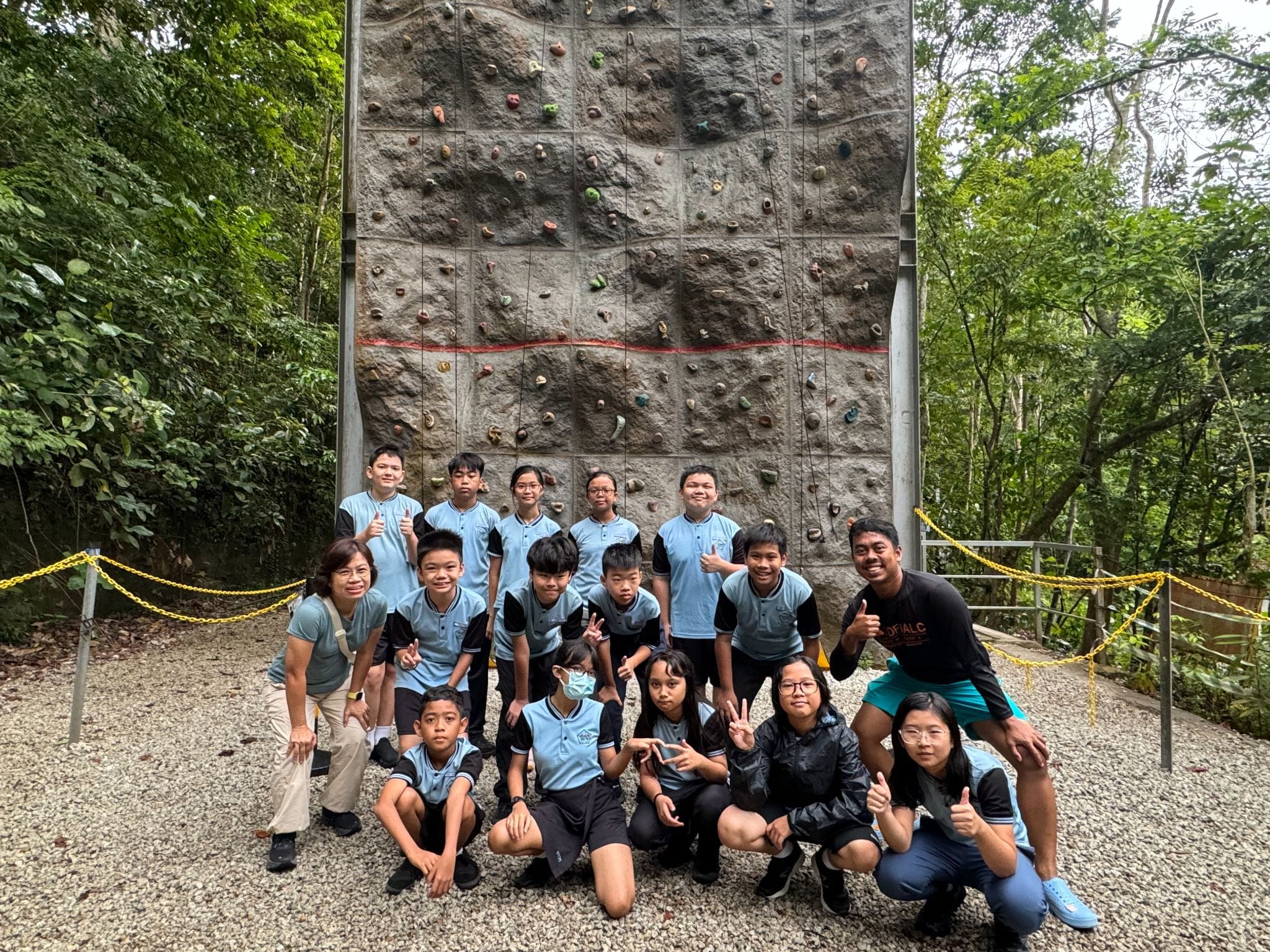 A group of students in blue uniforms posing in front of a tall outdoor rock climbing wall surrounded by trees.