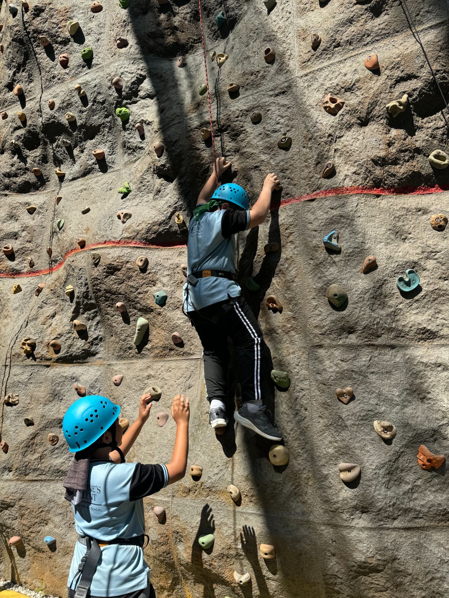 Two children wearing blue helmets on a rock climbing wall, one climbing up while the other watches from below.