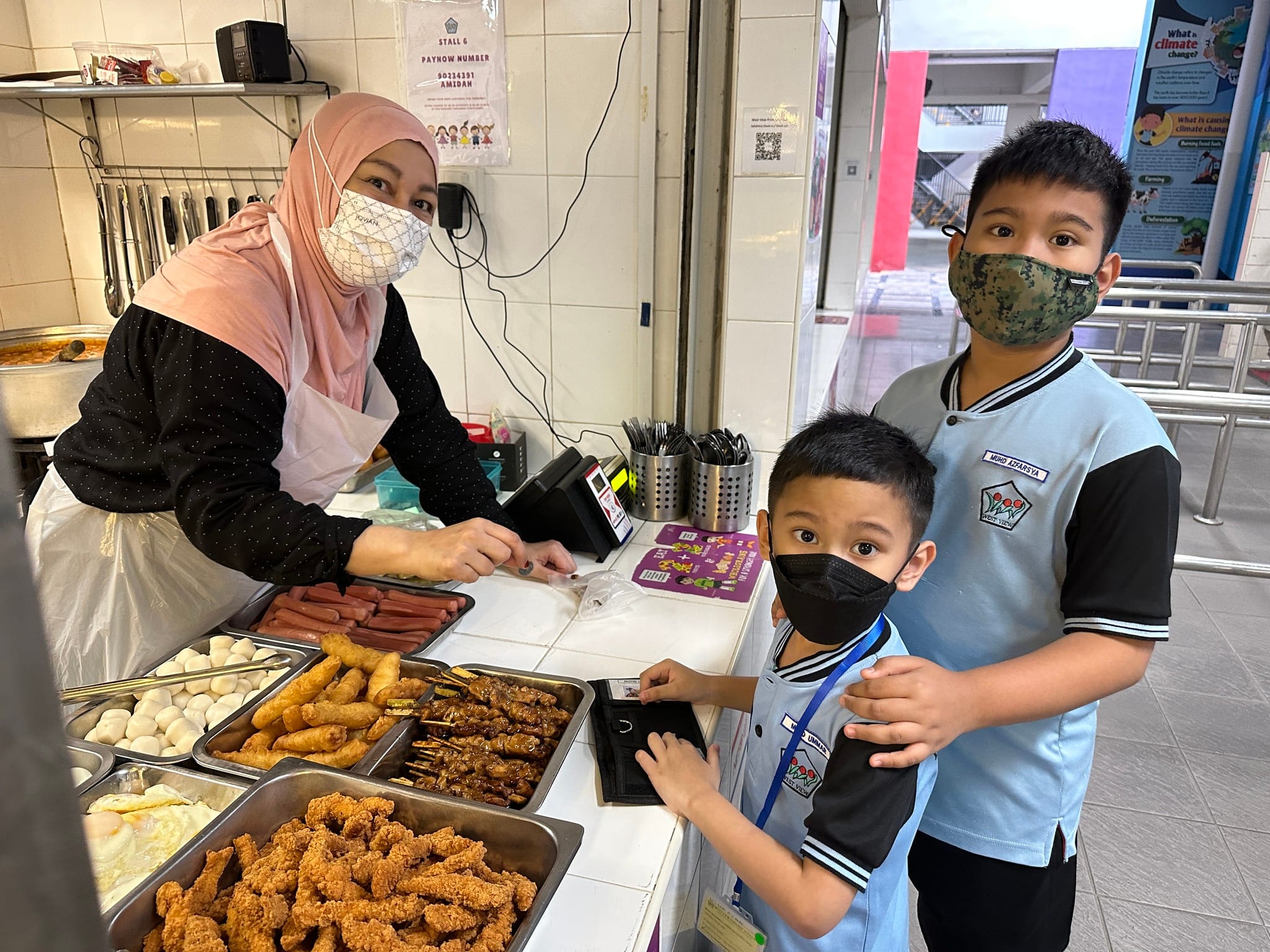 Two masked schoolboys buying food at a canteen stall from a woman vendor wearing a hijab and face mask.