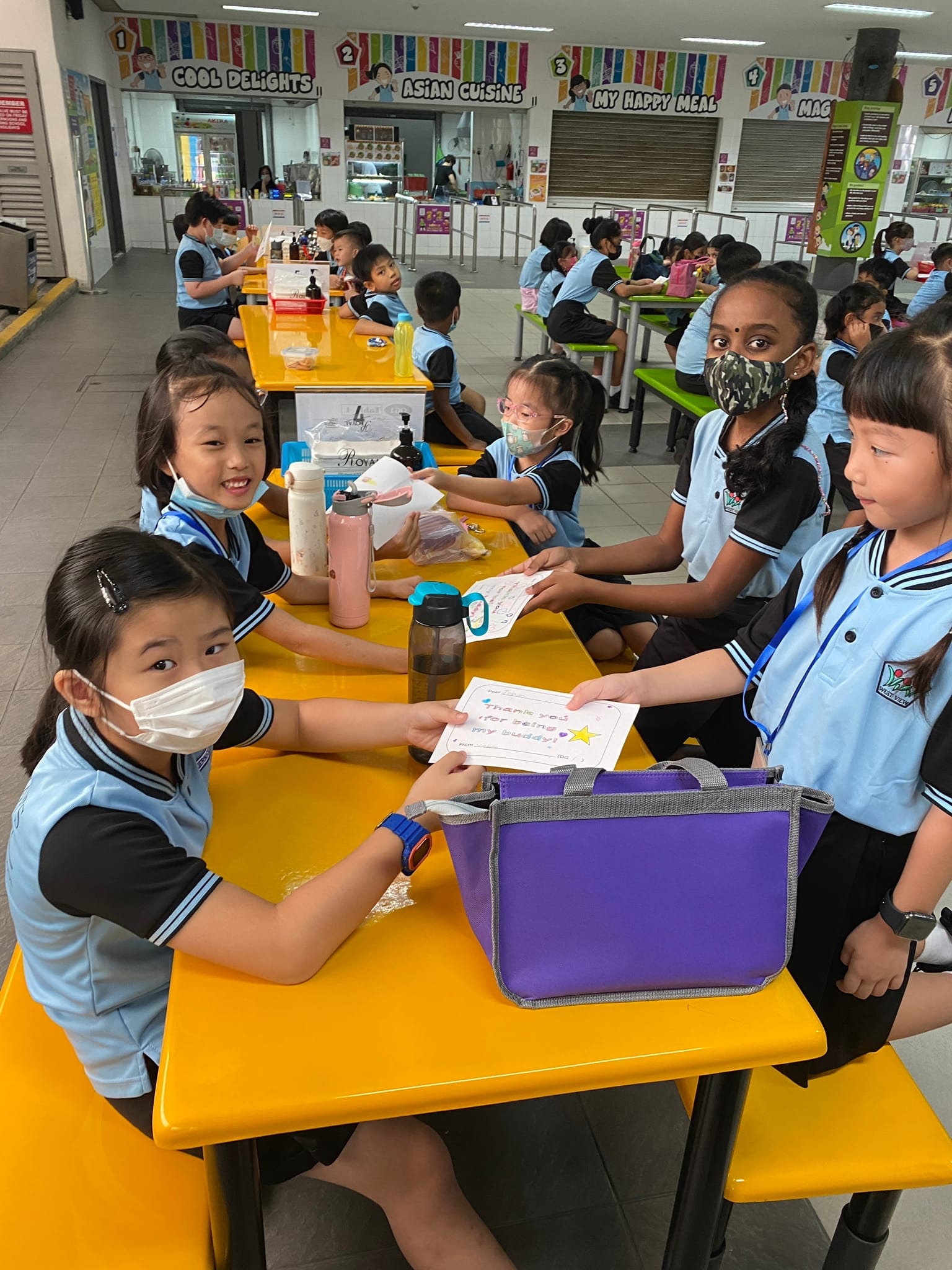 Young students in blue uniforms sit at yellow tables in a school canteen, exchanging a handwritten thank you card.