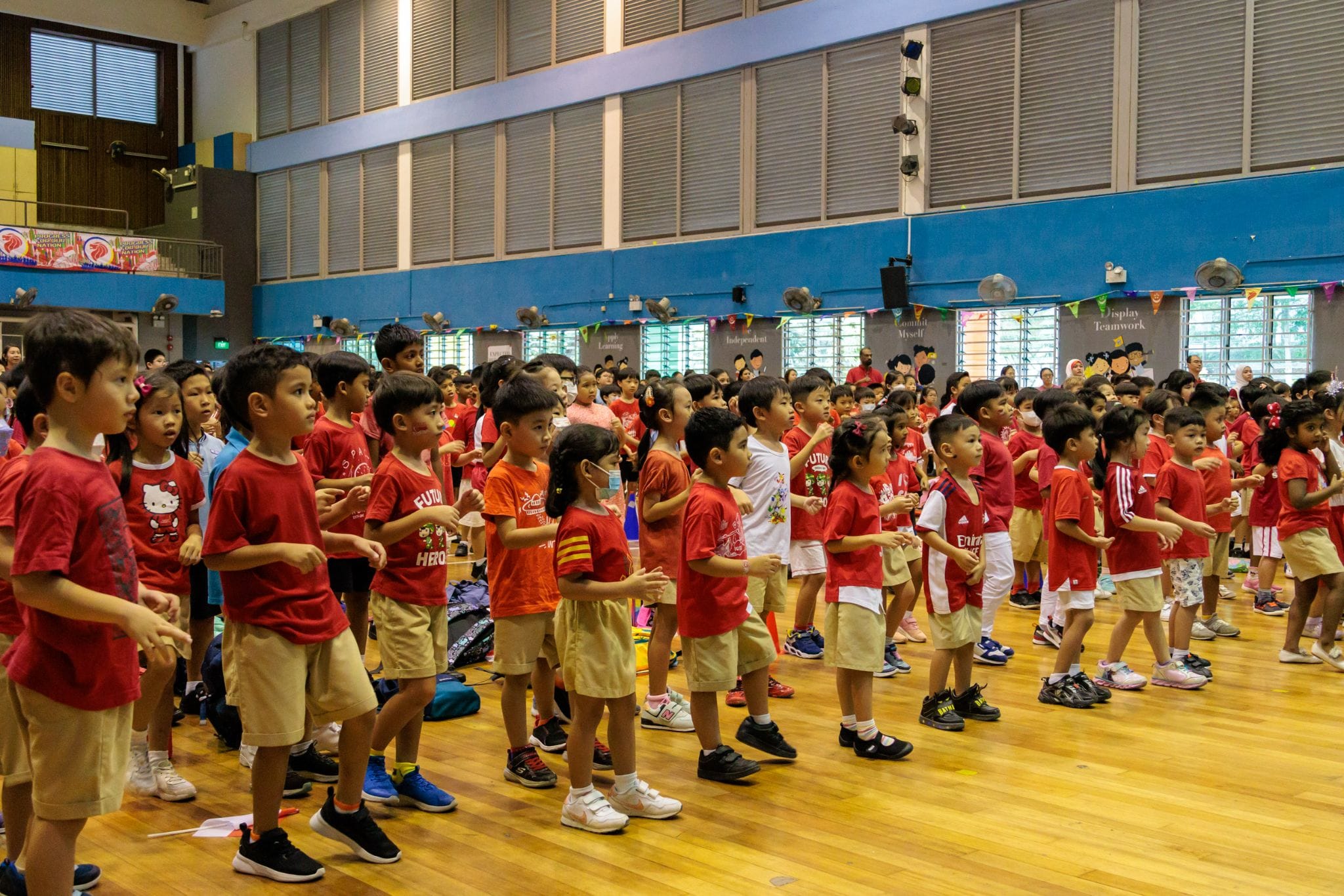 Large group of young children in red shirts and khaki shorts standing in rows inside a school hall.