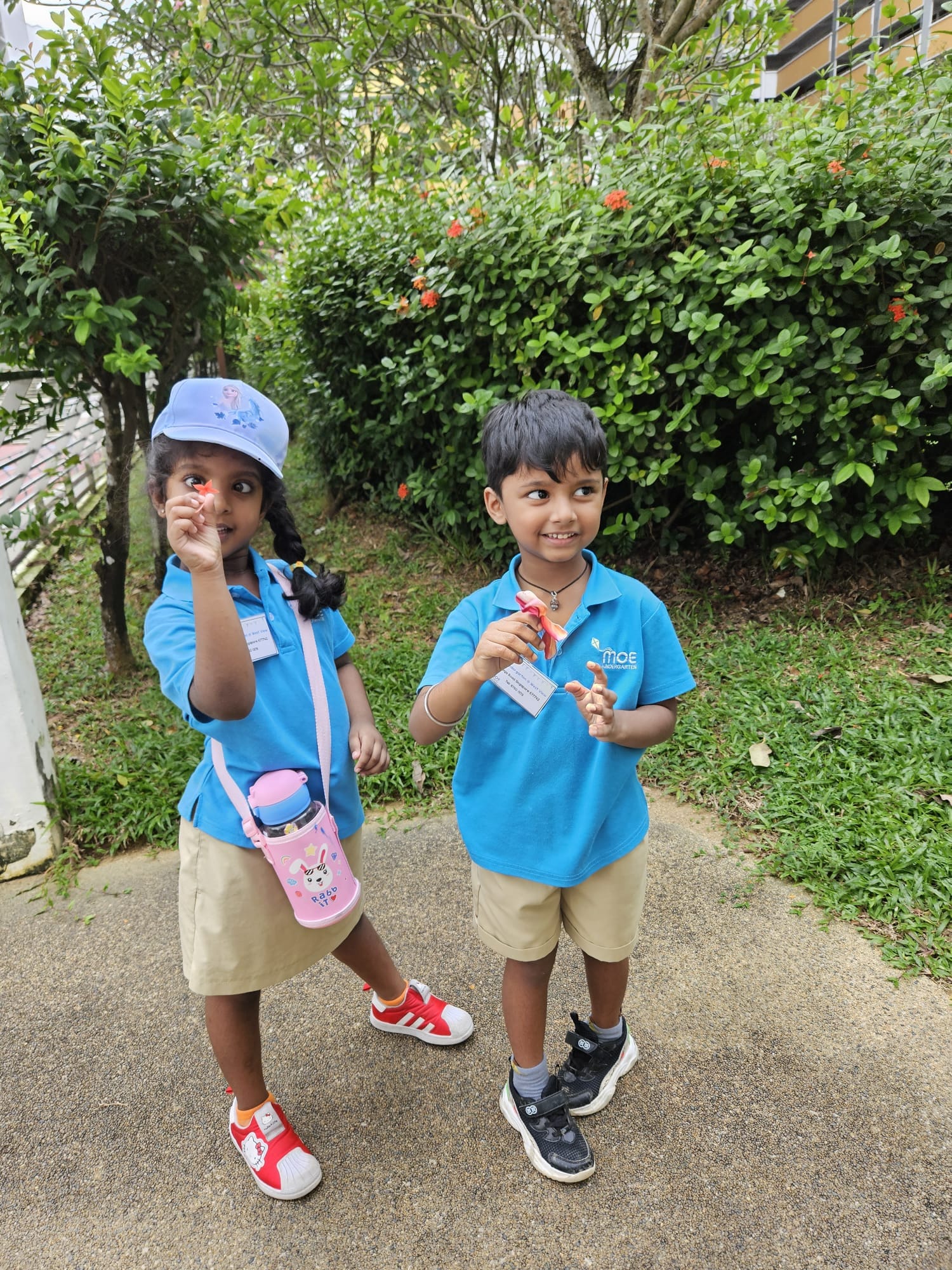 Two young children in blue school uniforms stand outdoors on a path, each holding small items, with green bushes behind them.