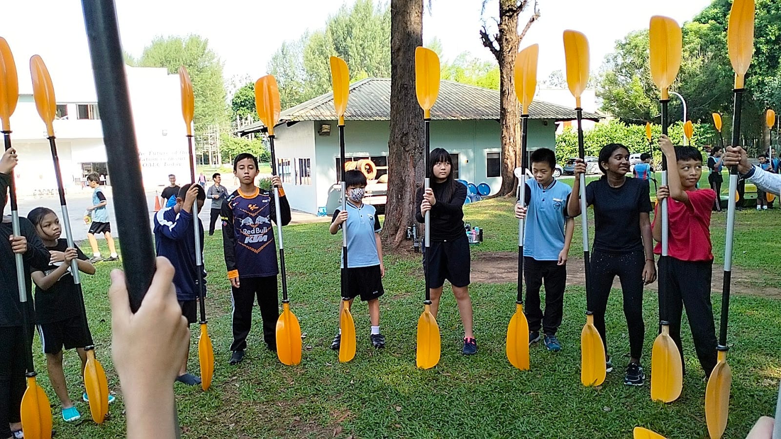 A group of children standing on grass outdoors, each holding a yellow kayak paddle upright during a training session.