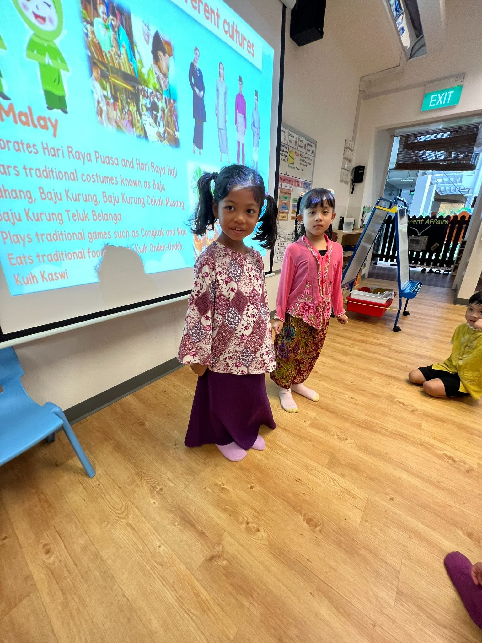 Two young girls in traditional Malay baju kurung stand in a classroom with a Malay culture slideshow projected behind them.