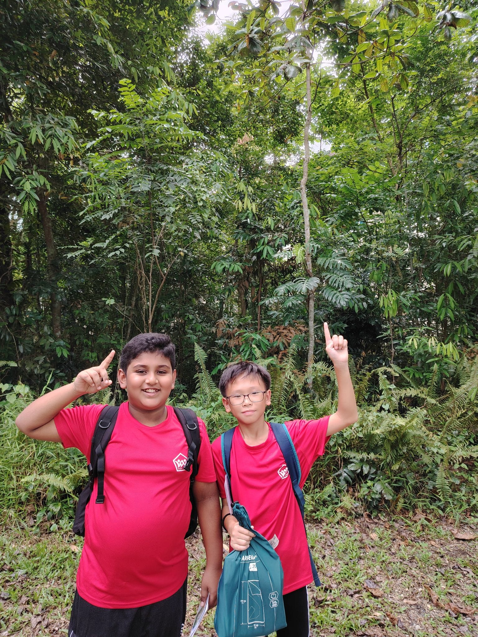 Two boys in red shirts with backpacks point upward while standing in front of dense green forest.