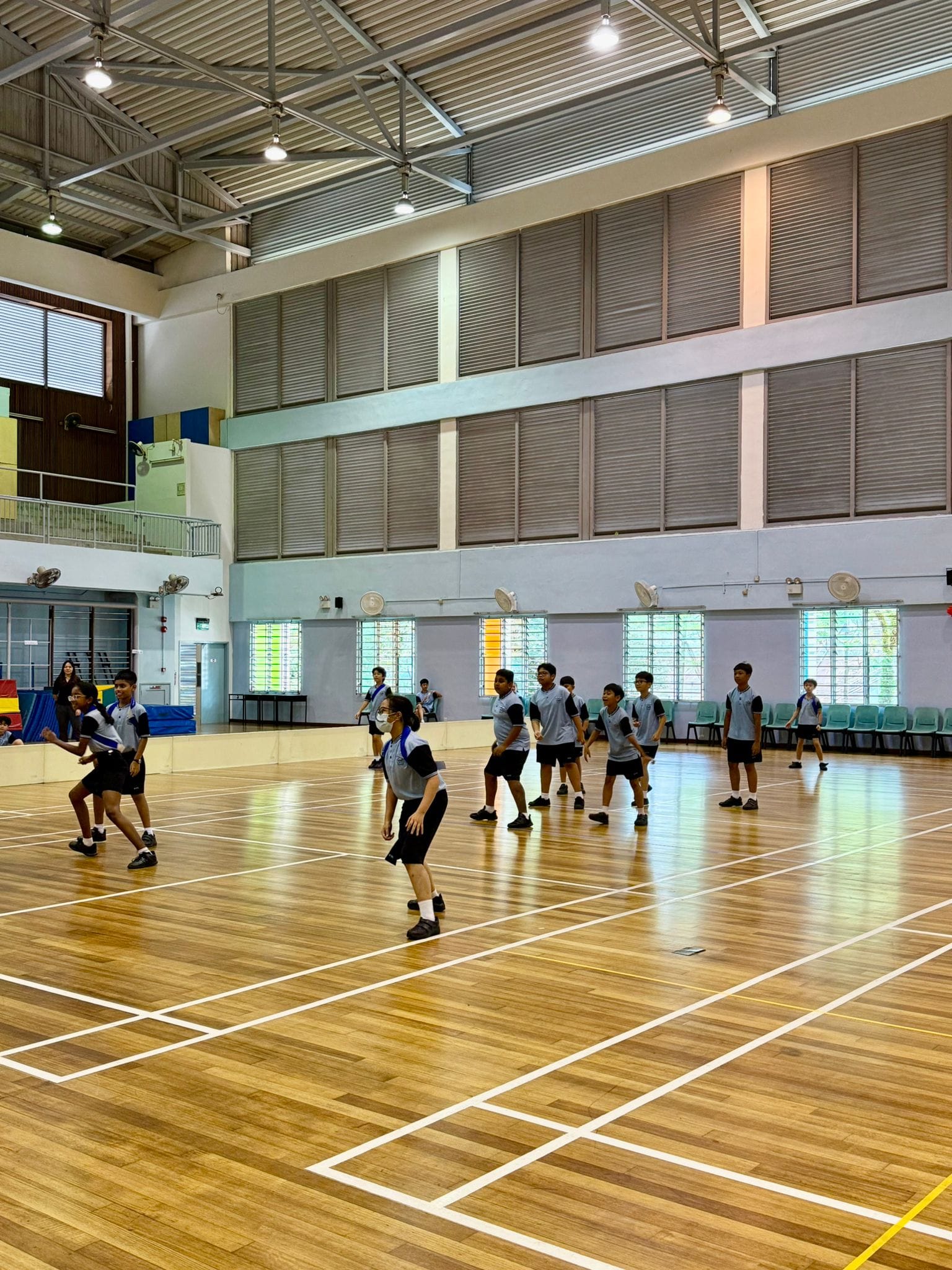 Students in gray uniforms exercise or practice drills on a wooden indoor sports hall court.