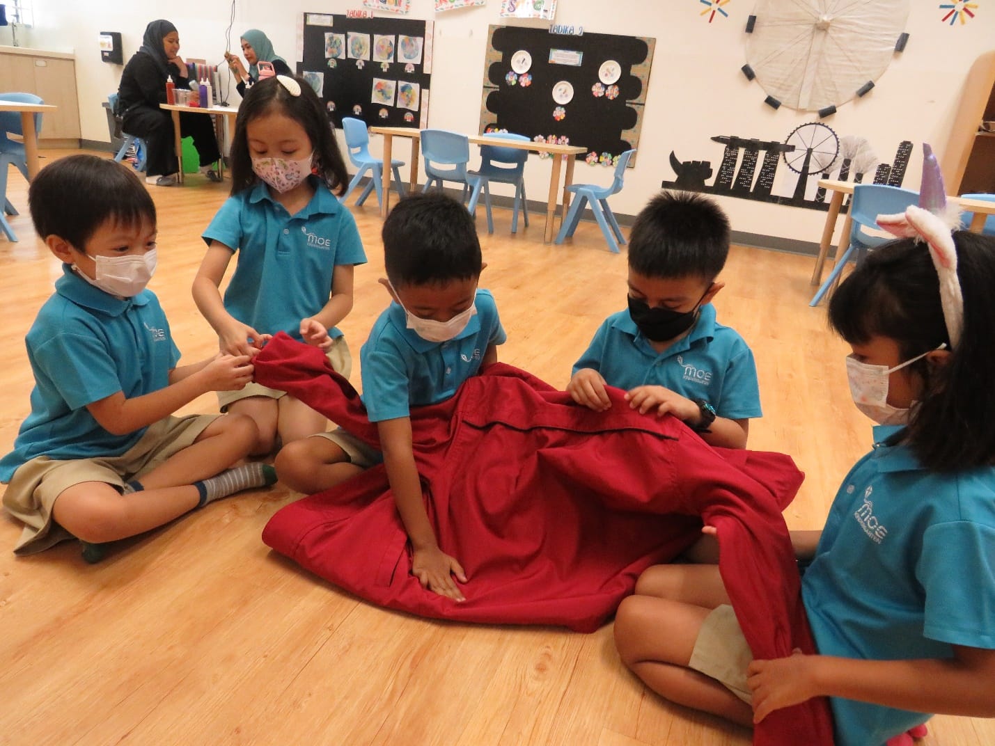 Four young masked children in teal uniforms sit on a classroom floor handling a large red fabric together.