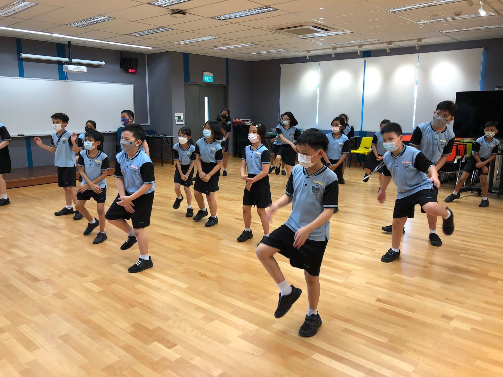 Children in blue and black school uniforms and face masks practice dance moves together in an indoor hall.