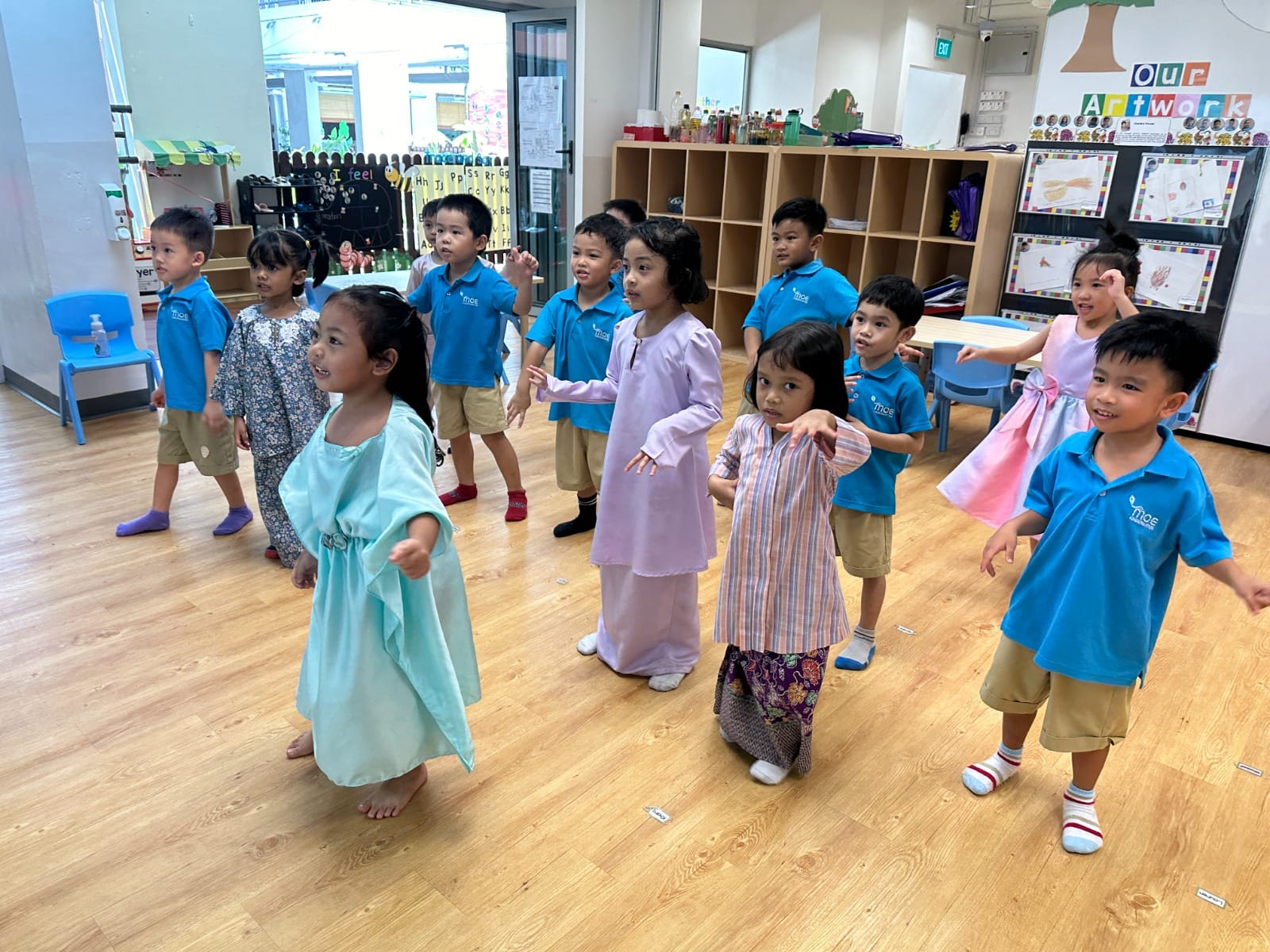 Young children in blue uniforms and traditional costumes dance together in a bright classroom with wooden floors.