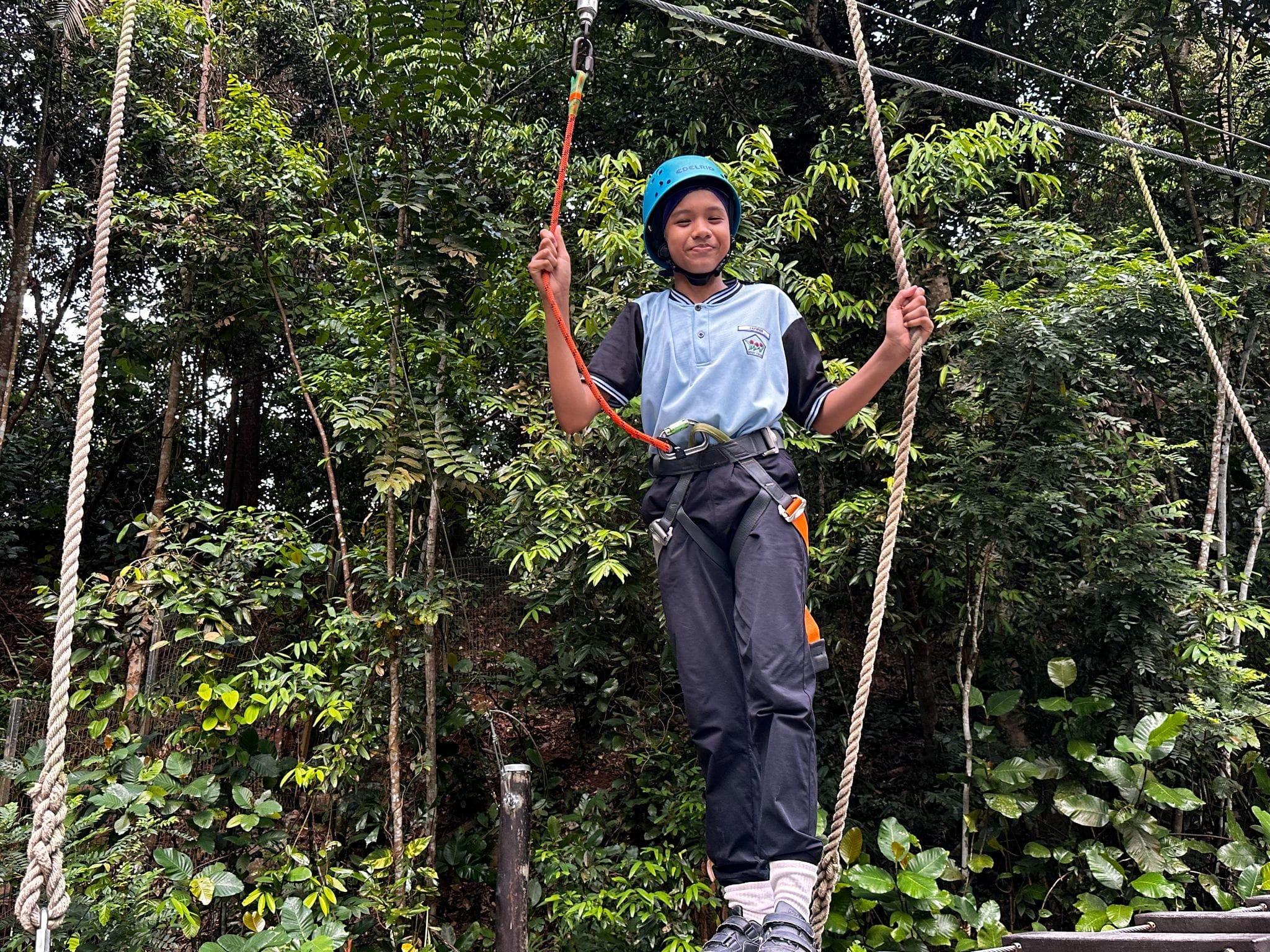 A child wearing a blue helmet and harness smiles while holding ropes on an outdoor high ropes course in a forest.