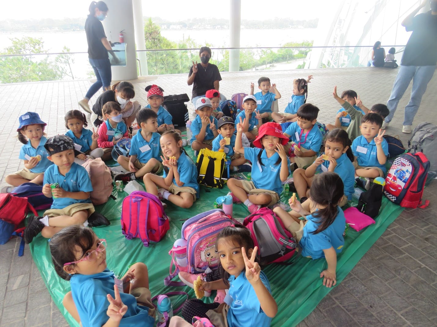 A large group of young children in matching blue uniforms sit on a green mat outdoors, waving peace signs with backpacks around them.