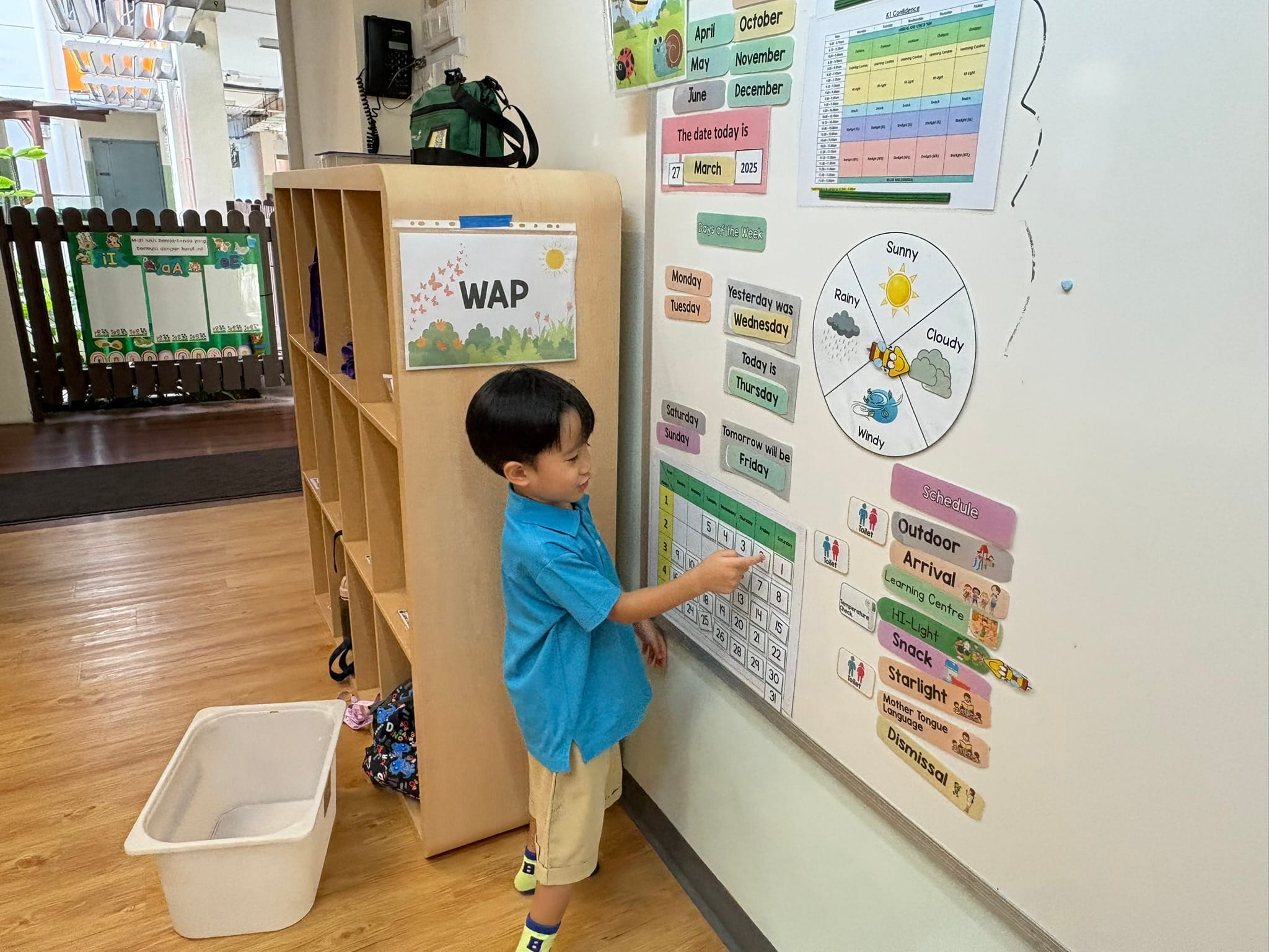 Young boy in blue shirt pointing at a classroom calendar wall display showing March 2025 with days and weather charts.
