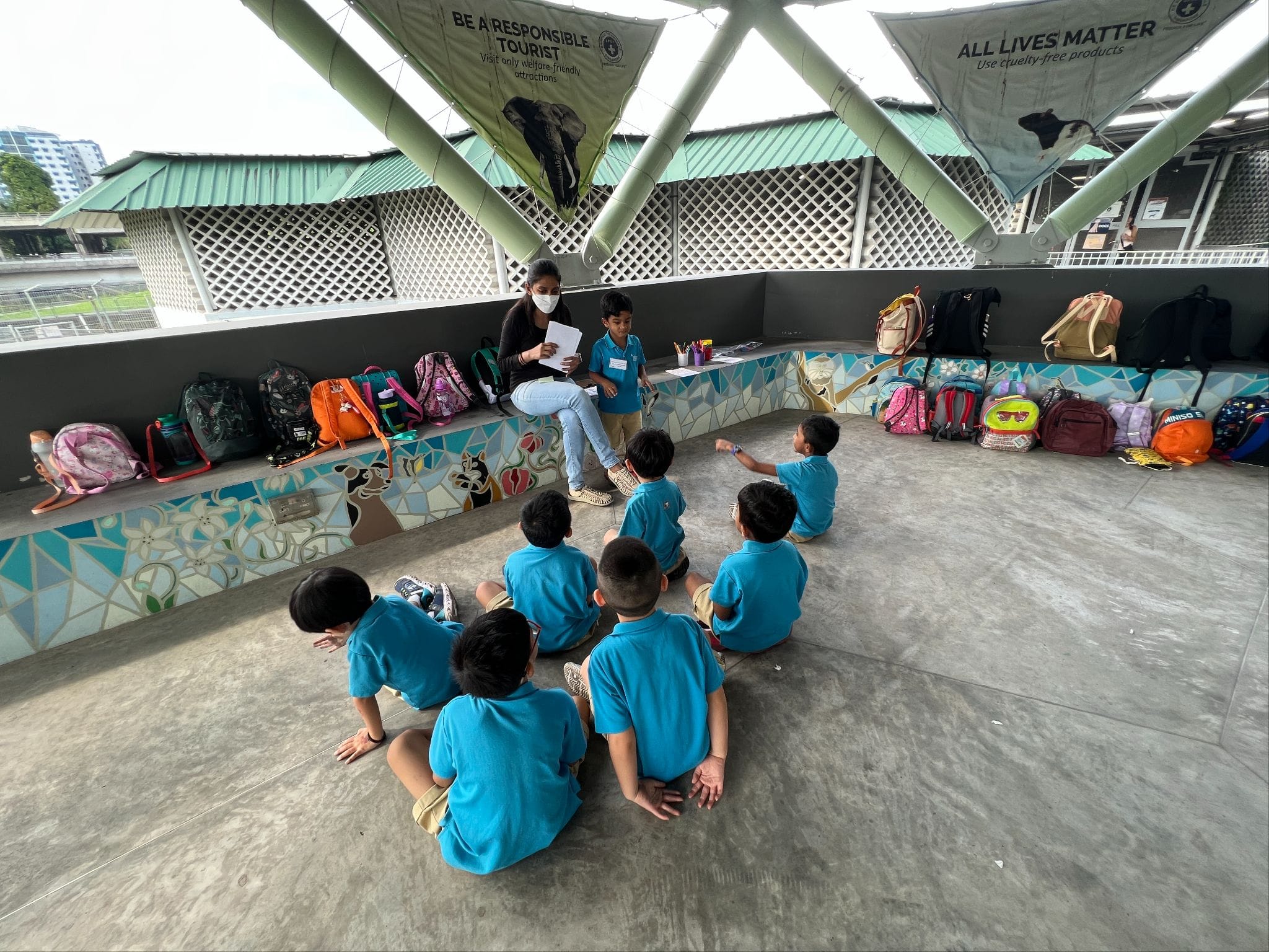 A masked adult sits on a mosaic bench reading to a group of young children in blue uniforms seated on the floor.