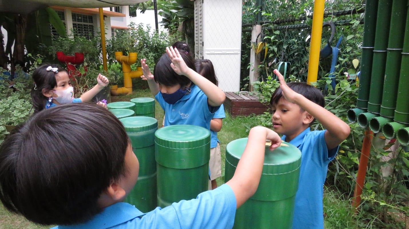 Children in blue uniforms play outdoors with green cylindrical drums in a garden setting.