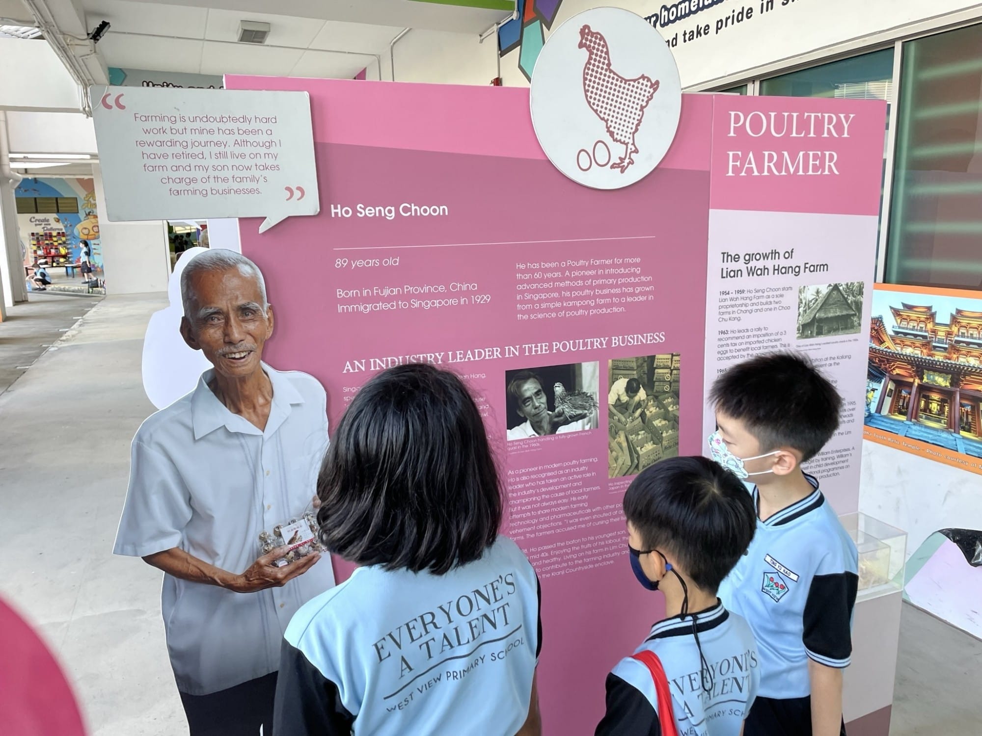 Three primary school students view a pink exhibition board about poultry farmer Ho Seng Choon beside his life-size cutout.