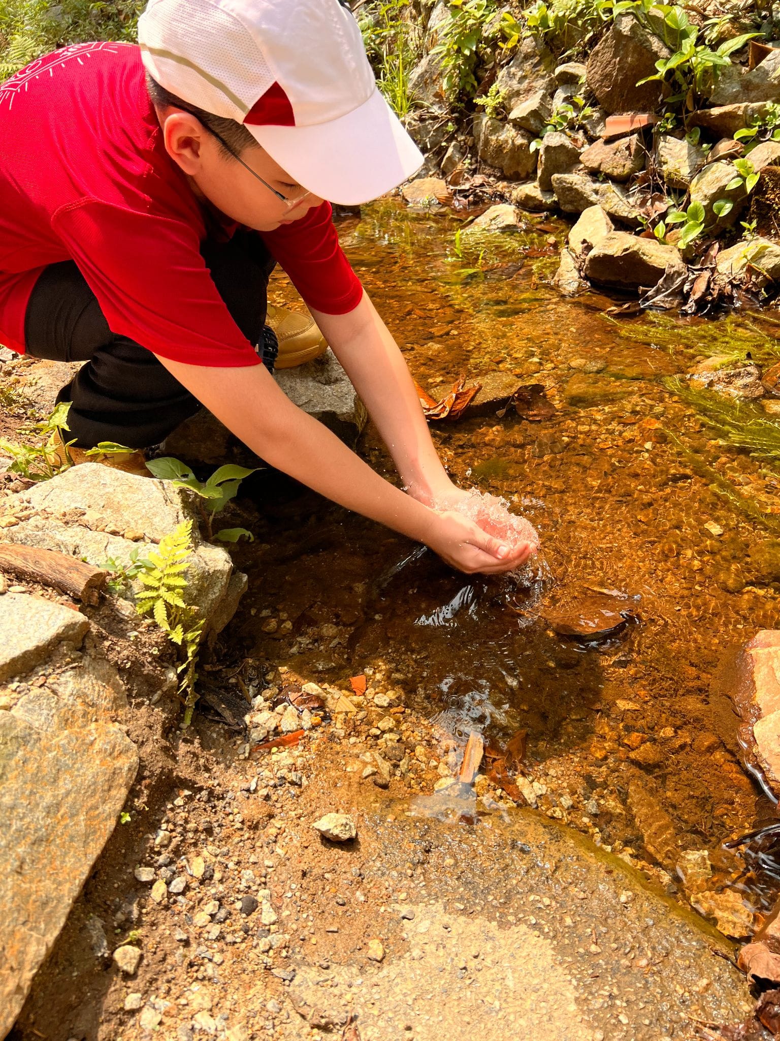 A child in a red shirt and white cap crouches beside a shallow rocky stream, scooping water with cupped hands.