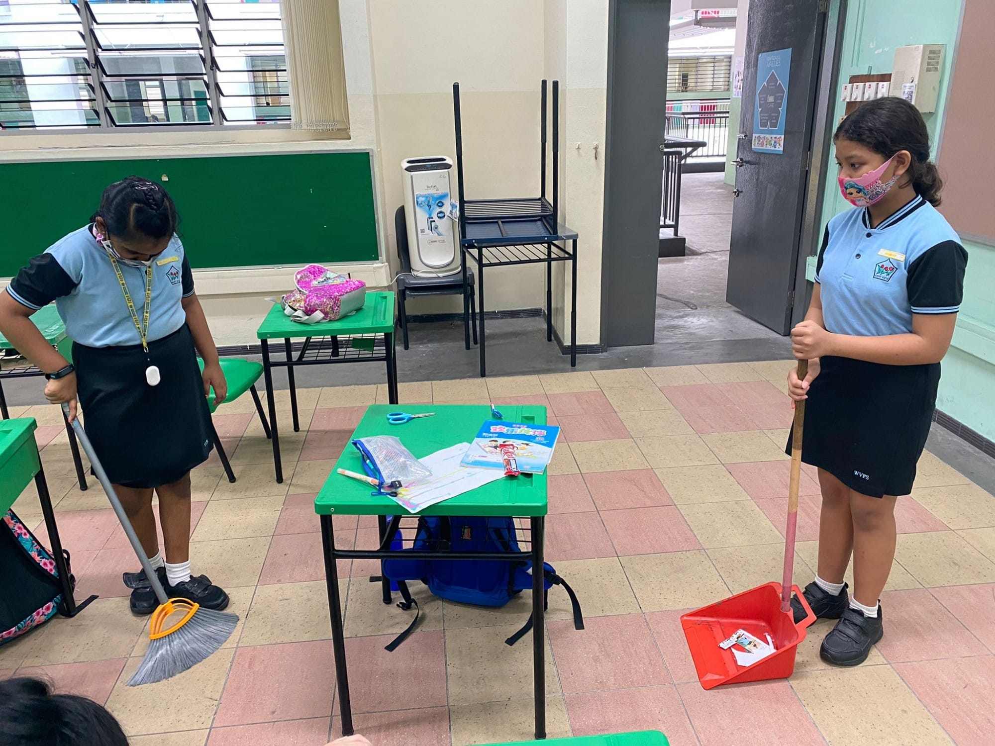 Two masked schoolgirls in blue uniforms sweep a classroom floor with a broom and dustpan.