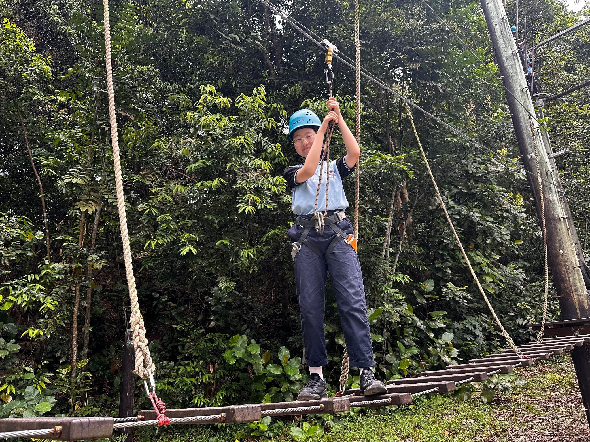 A child wearing a blue helmet and harness walks across a rope bridge on an outdoor ropes course surrounded by trees.