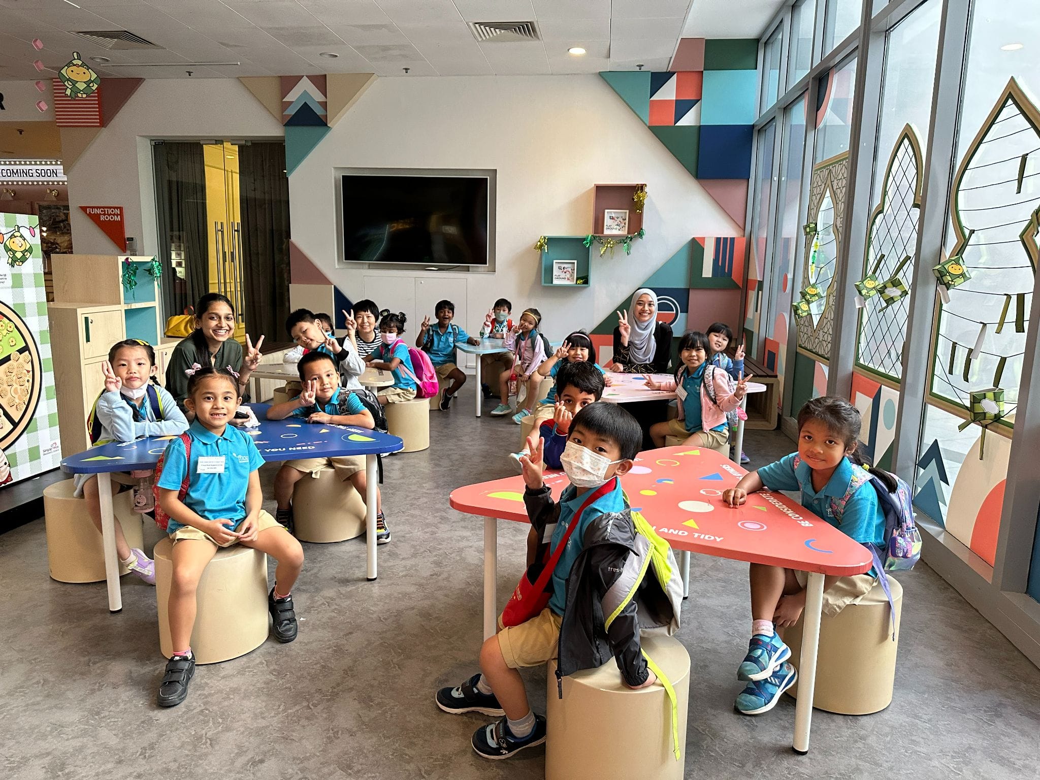 Young children in blue uniforms sit at colorful tables in a bright room, smiling and making peace signs.