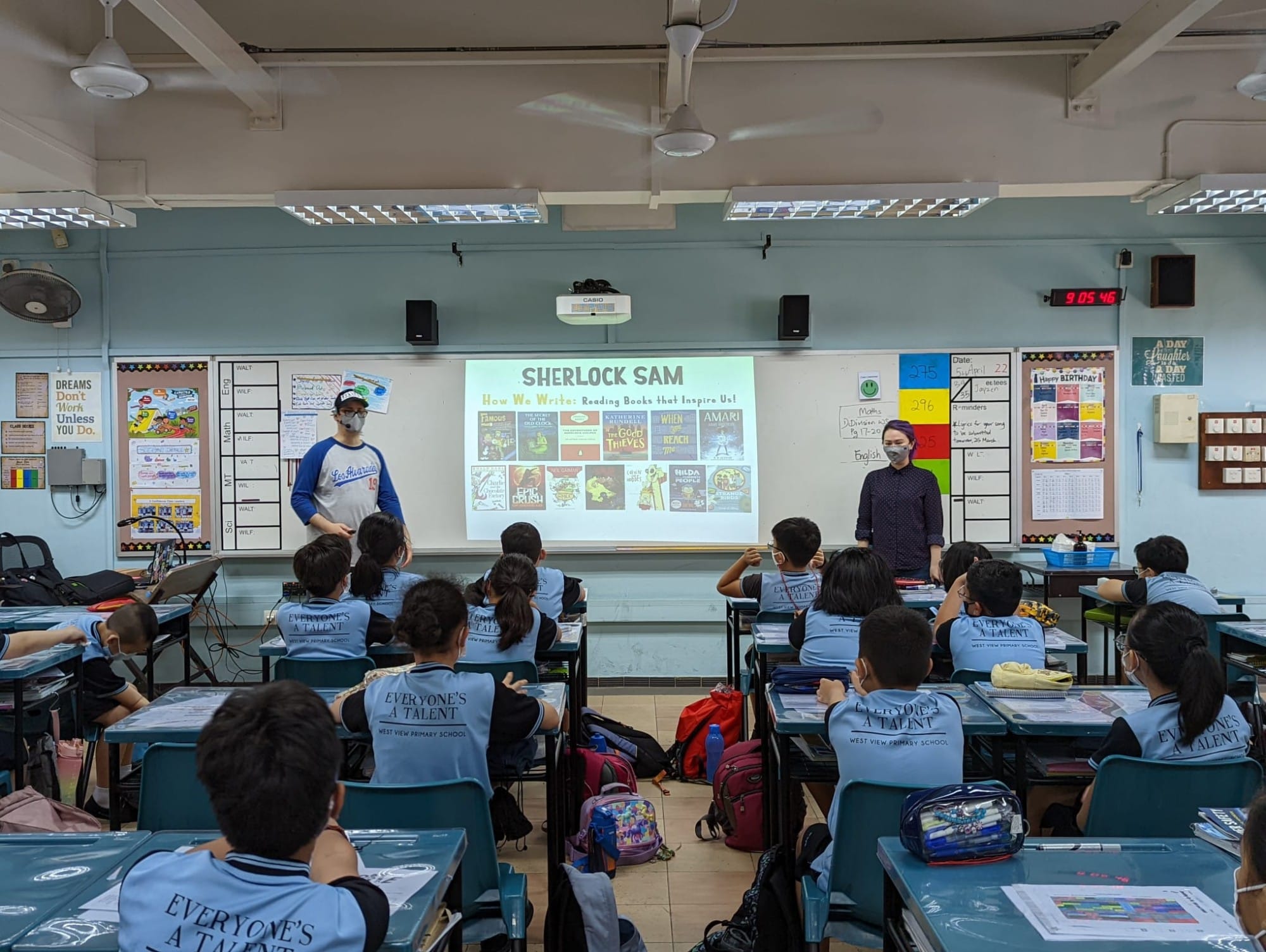 Two teachers present a Sherlock Sam reading lesson to uniformed primary school students seated at desks.