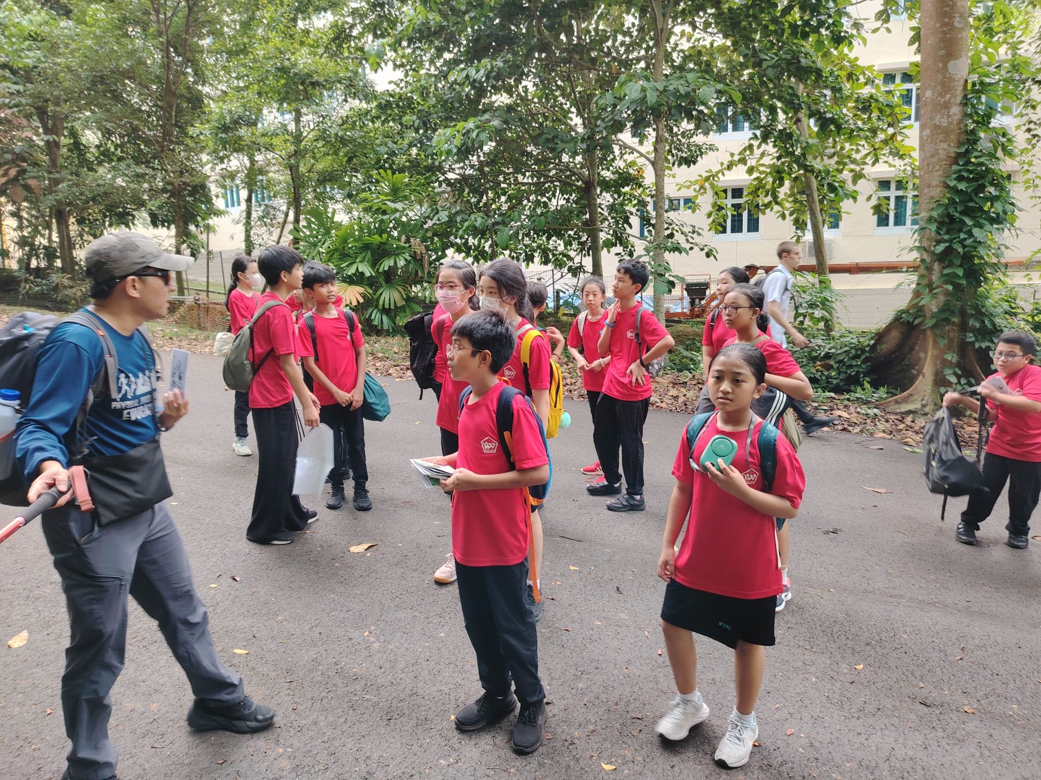 A group of children in red shirts with backpacks stand outdoors near trees and a building, listening to an adult instructor.