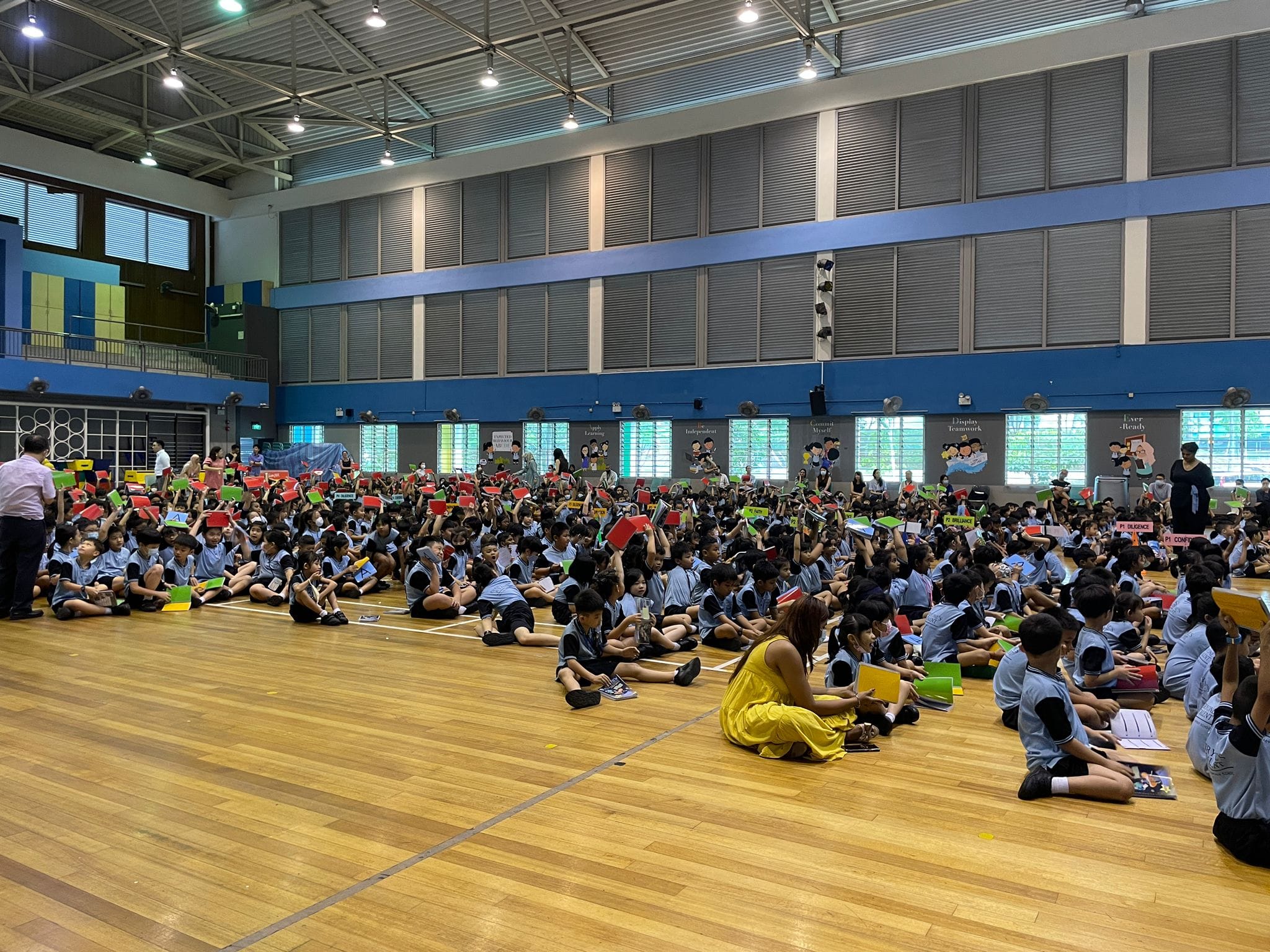 Large group of school children in uniforms sitting on a gymnasium floor holding colorful red, green, and yellow cards.