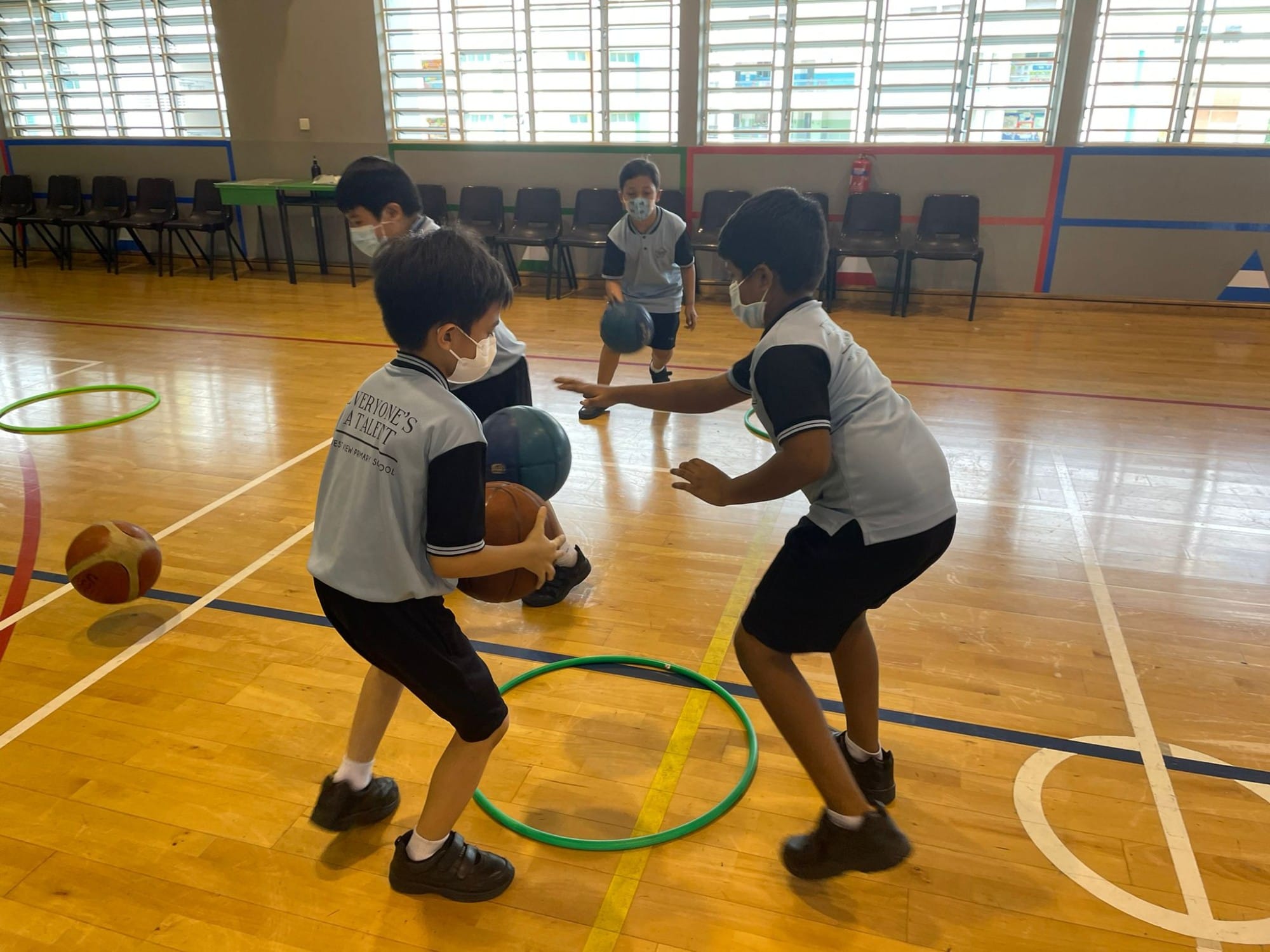 Four masked school children in uniforms play with balls and hula hoops on an indoor gymnasium court.
