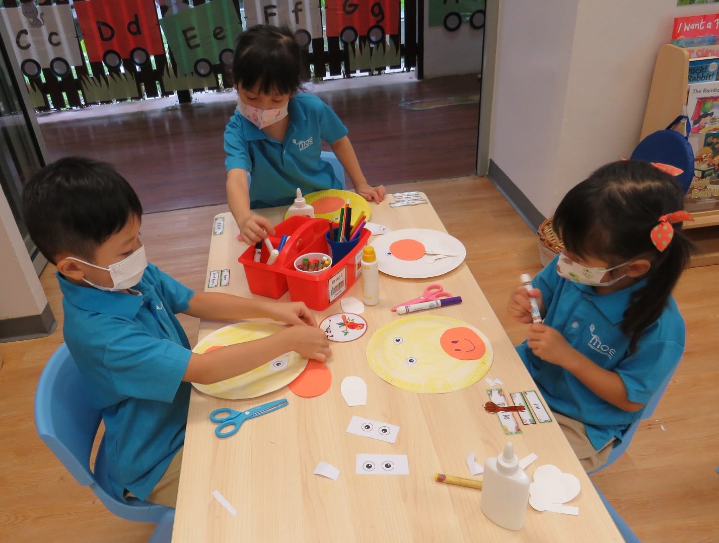 Three masked children in blue uniforms do a paper plate craft with scissors, glue, and colored paper at a classroom table.