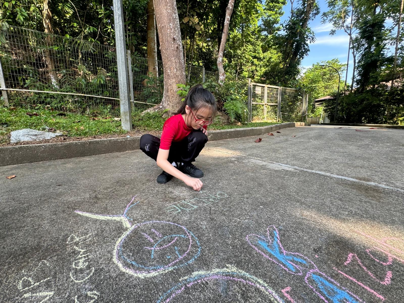 A young girl in a red shirt crouches on pavement drawing colorful chalk artwork outdoors near trees.