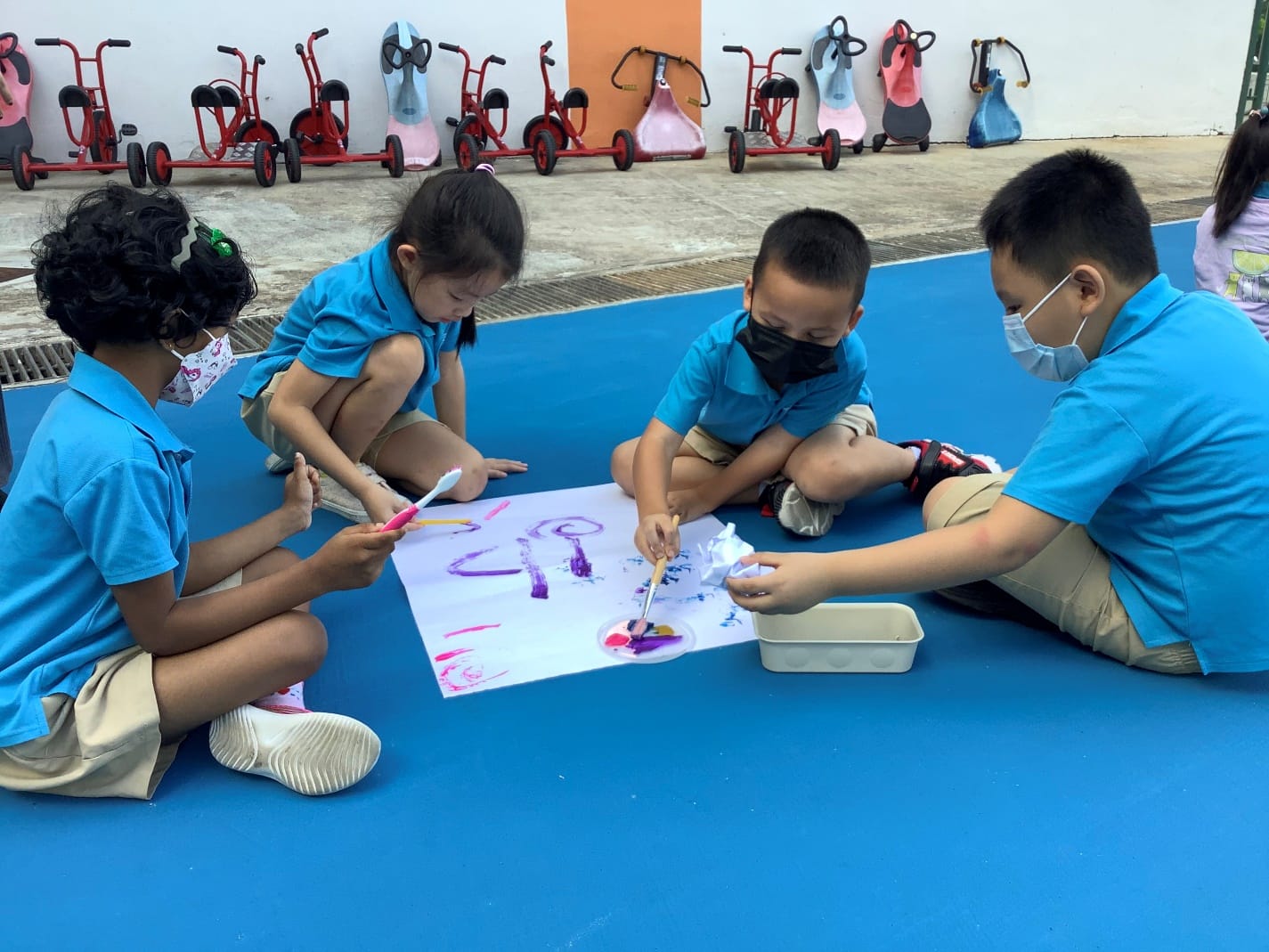 Four young children in blue uniforms sit on a blue floor painting together on a large white sheet outdoors.