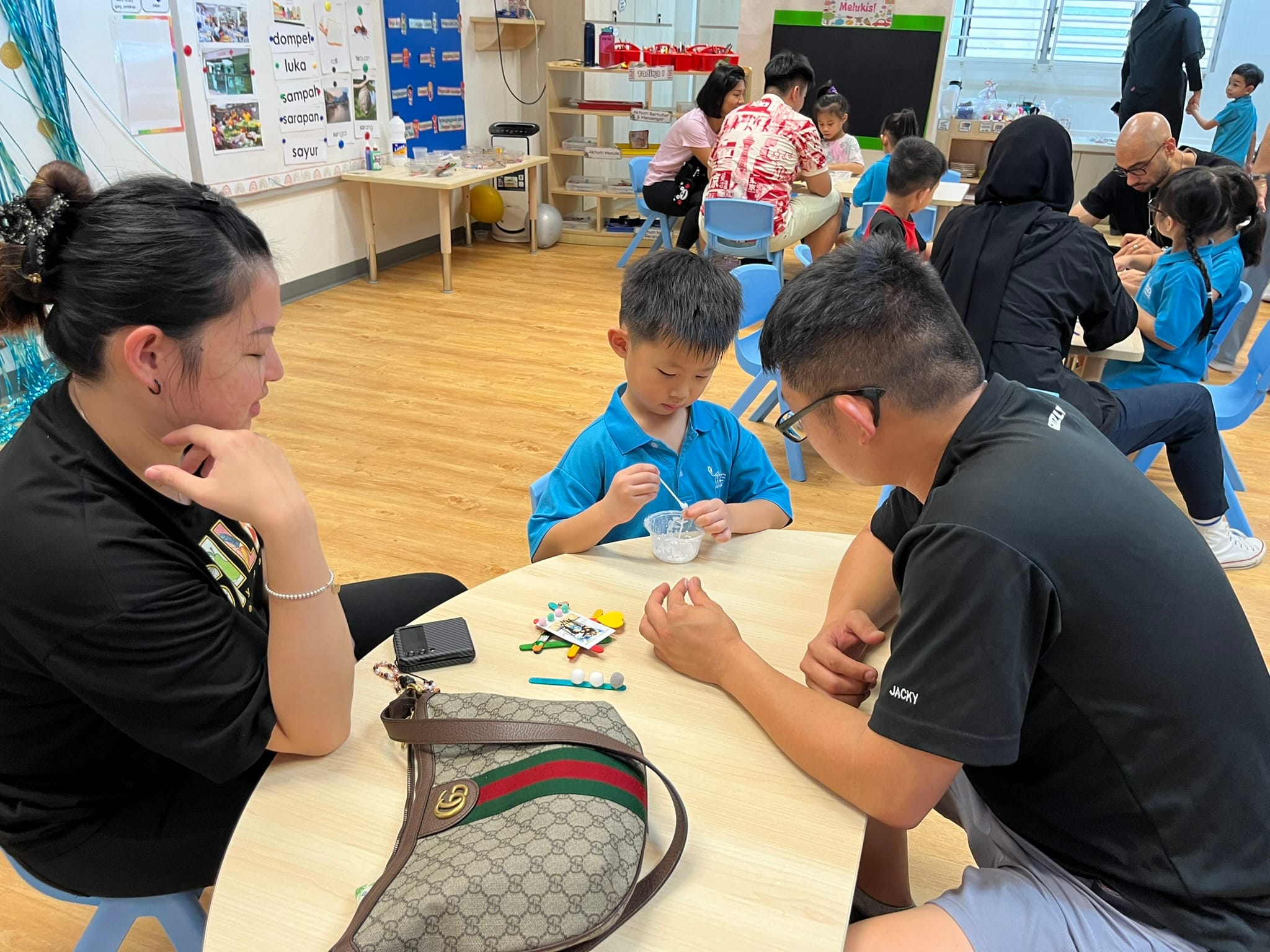 A young boy in a blue shirt does a craft activity at a table with two adults in a busy classroom.