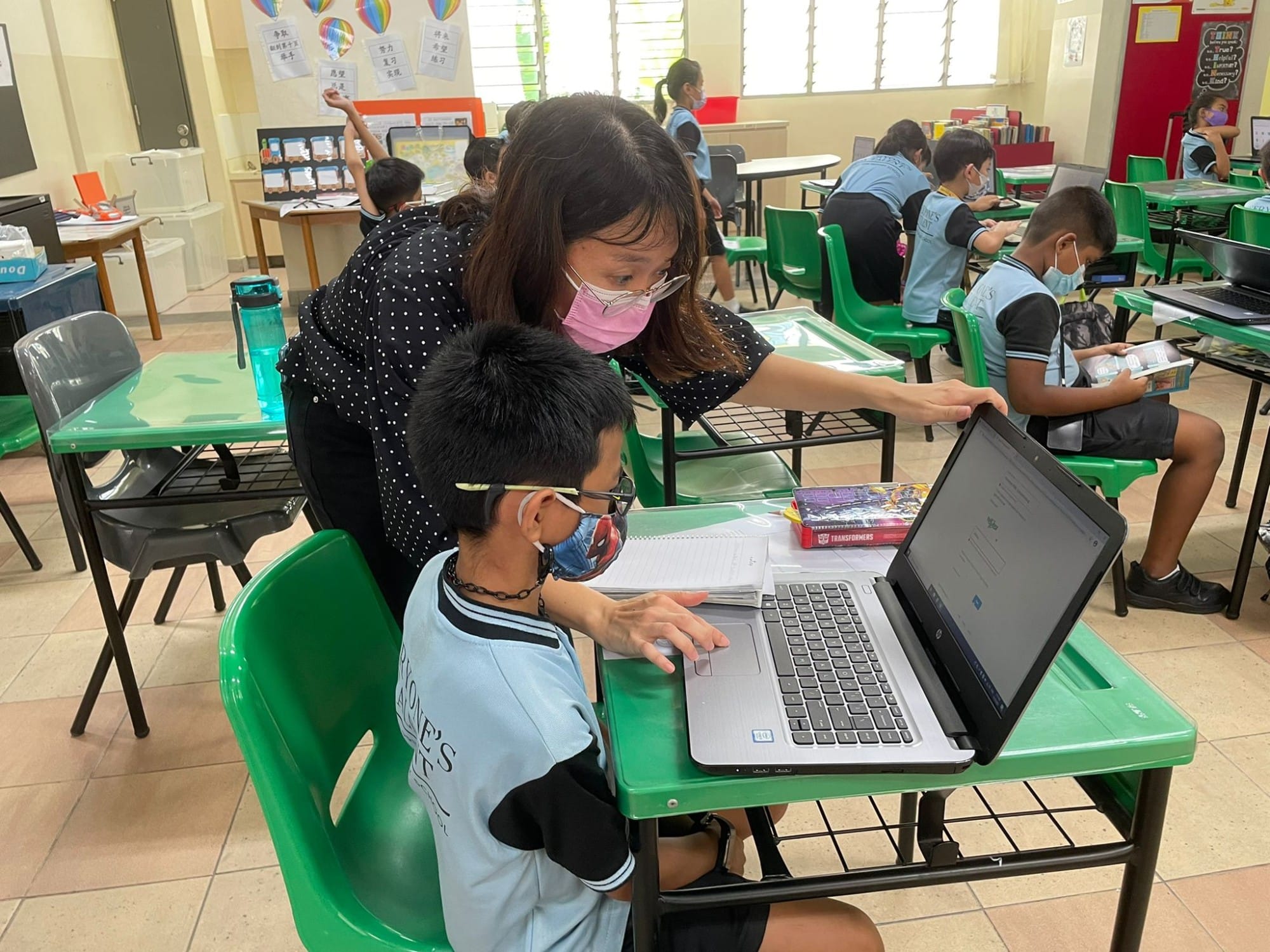 A masked teacher leans over to help a young masked student working on a laptop at a green desk in a classroom.