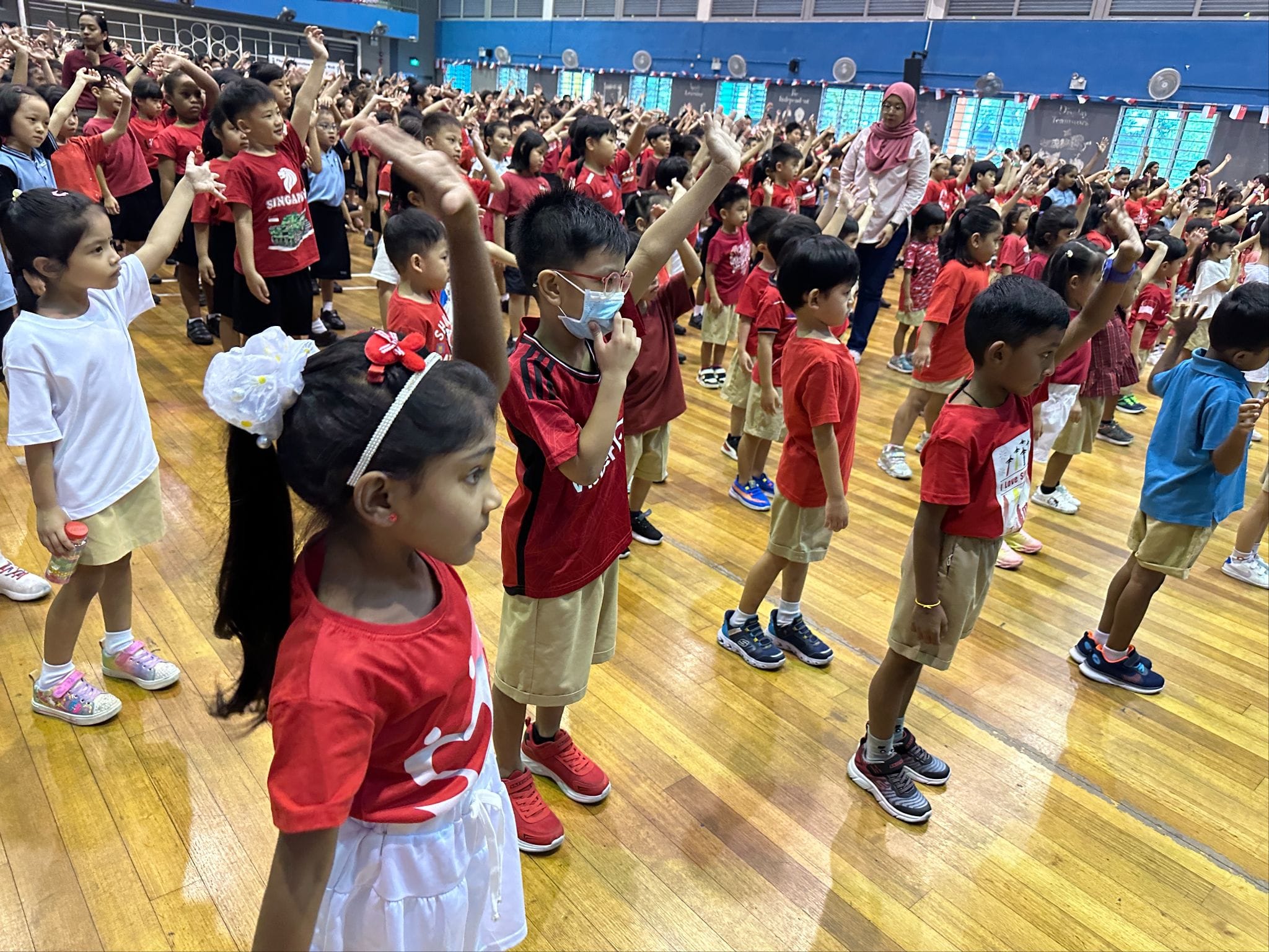 Large group of young children in red shirts raising hands during a school event in an indoor gymnasium.