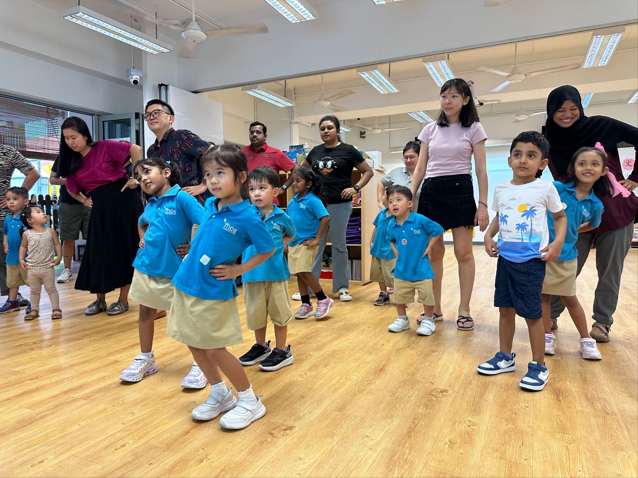 Young children in blue and khaki uniforms perform a dance or activity indoors while adults watch nearby.