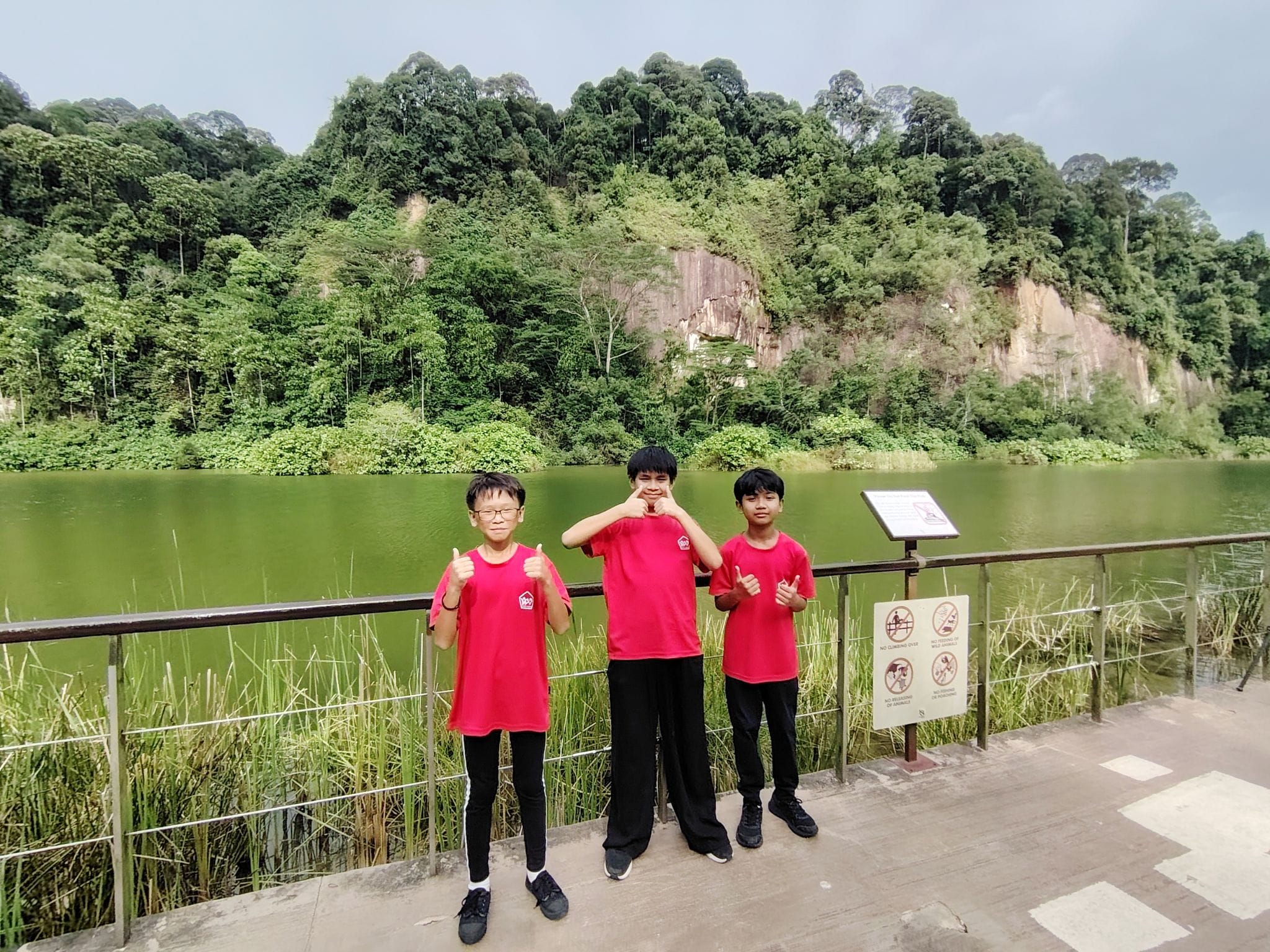 Three boys in red shirts pose by a railing in front of a green lake with a rocky forested cliff behind them.