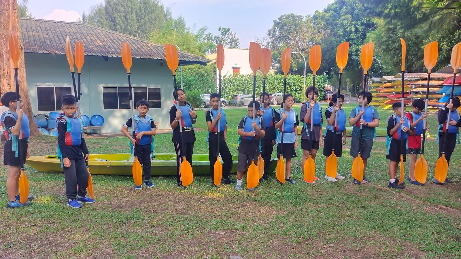 A group of children in blue life vests stand in a line outdoors, each holding an orange kayak paddle upright.