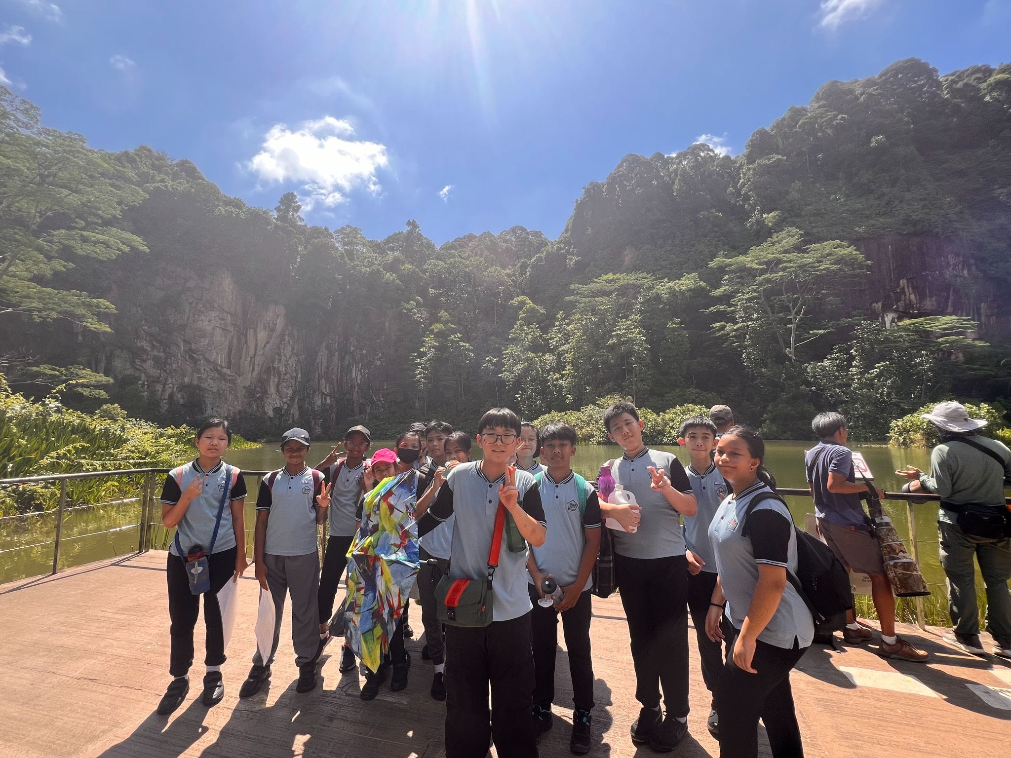 A group of students in light blue uniforms pose on a platform near a lake with tall forested limestone cliffs behind them.